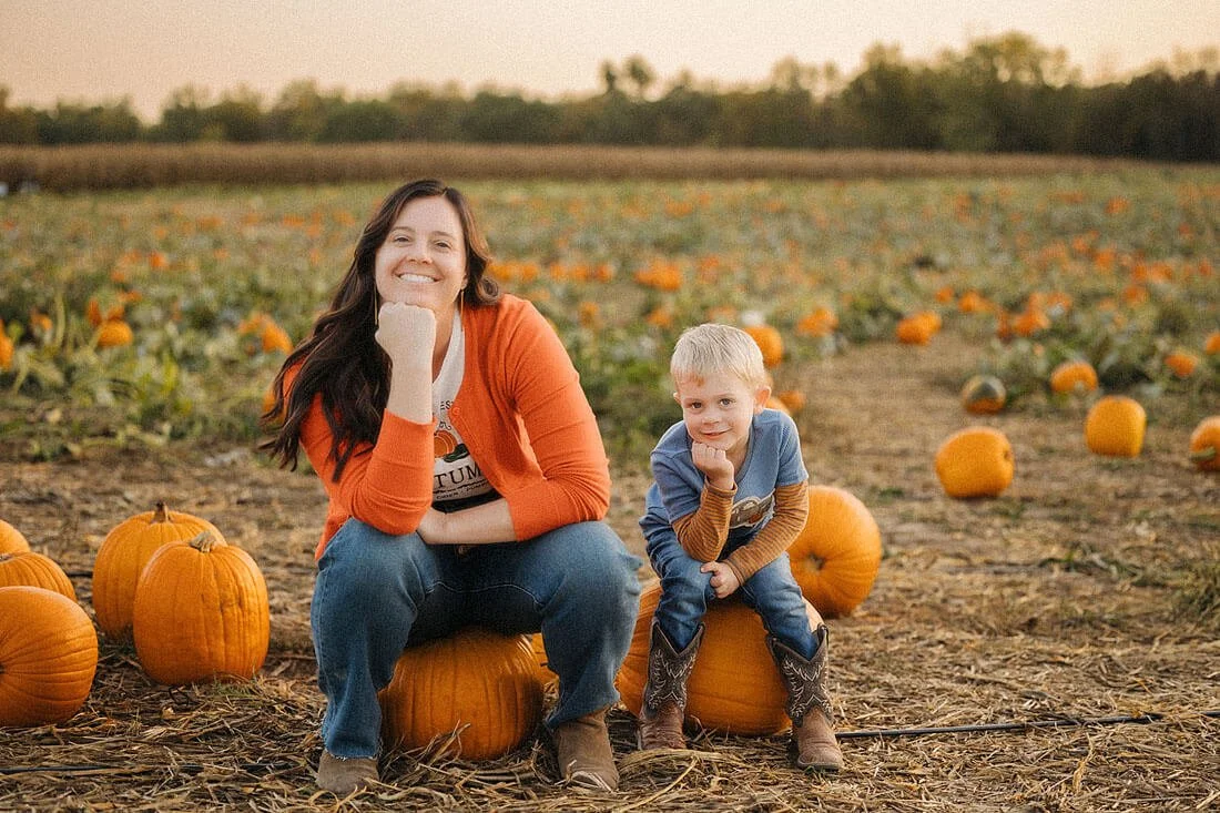 Mother and young son sit on pumpkins at Dull’s Pumpkin Harvest, smiling with their chins resting on their hands in matching poses