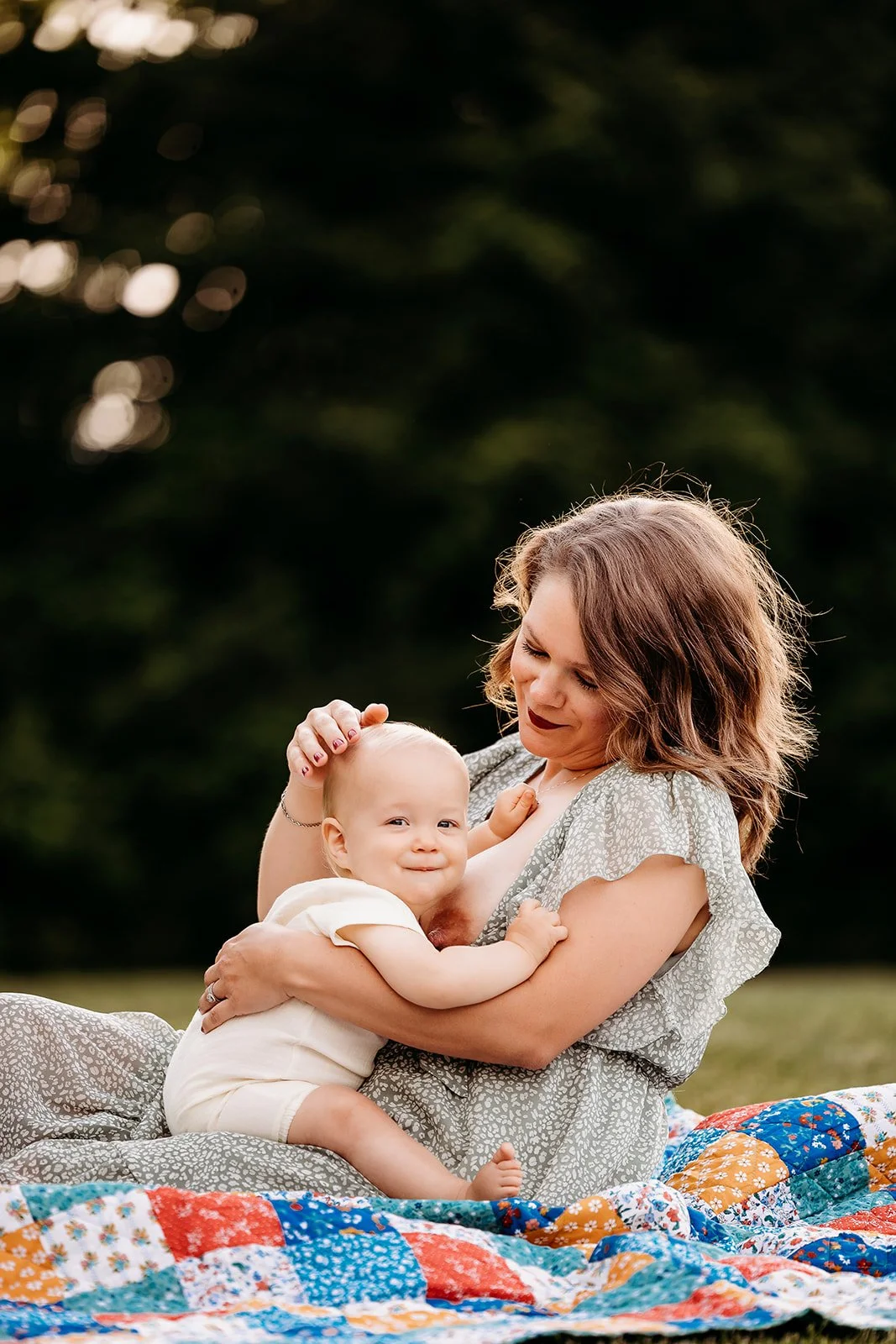 A woman and a baby sitting on a colorful quilt outdoors, with the woman holding the baby in her arms and both smiling.