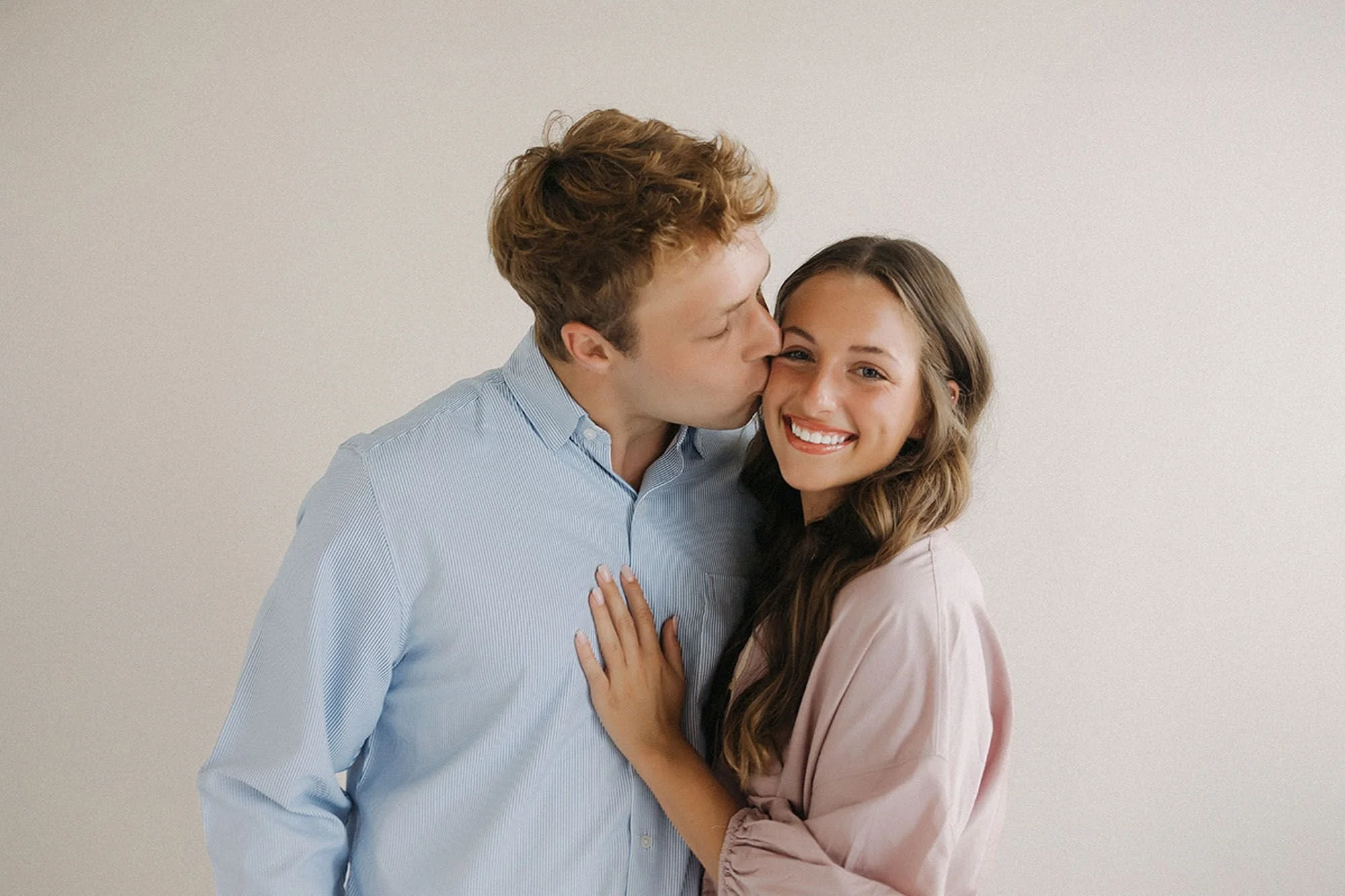 A curly-haired man in a light blue shirt kisses his smiling partner on the cheek while she rests her hand on his chest, both glowing warmly against a soft neutral backdrop