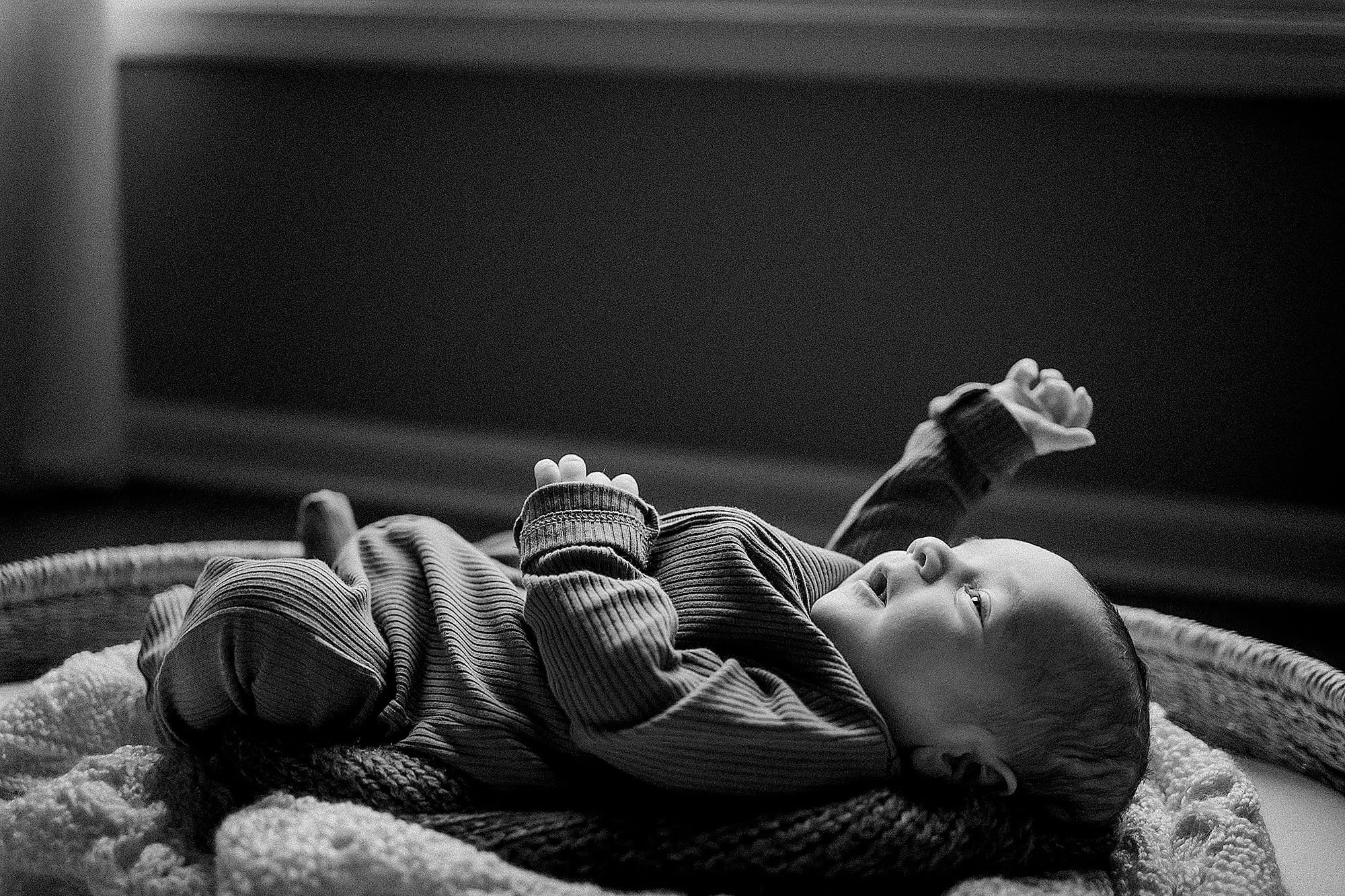 A baby lying on a textured blanket, looking up with one arm raised in a darkened room.