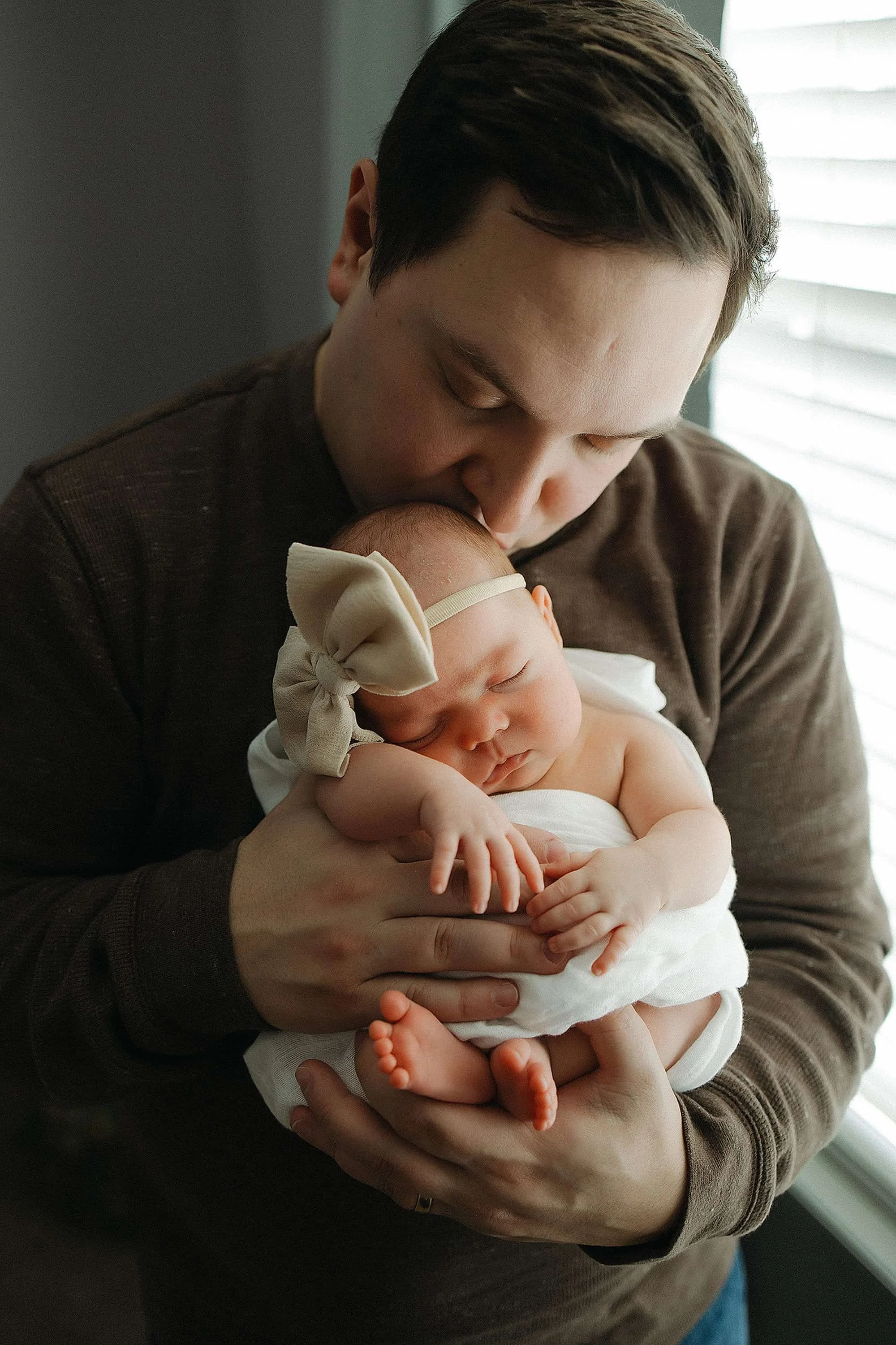 A man holding a sleeping baby girl, who is wearing a beige bow headband and white outfit, near a window with blinds.
