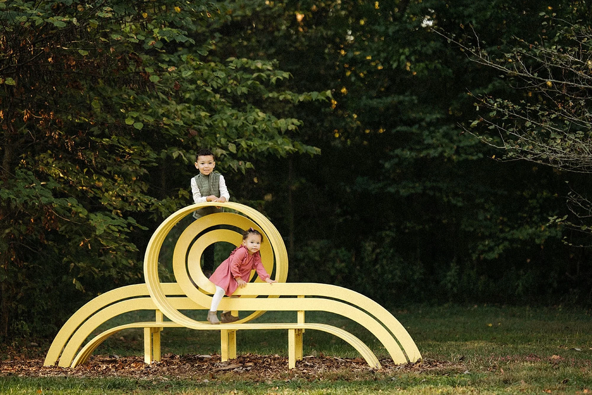 Young siblings climbing a bright yellow spiral sculpture bench surrounded by evergreen trees at Virginia B. Fairbanks Art & Nature Park, family photos featuring playful moments with public art