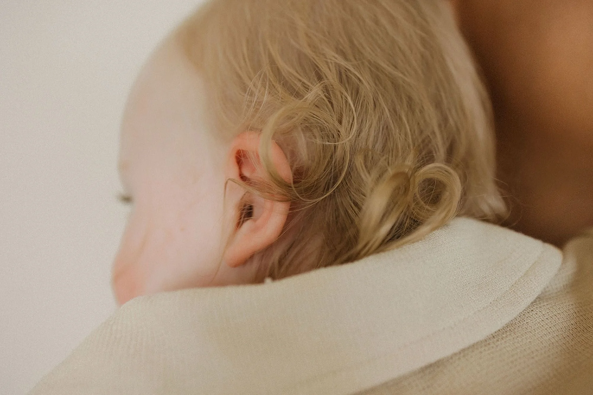 Close-up profile shot of a blond toddler's soft curls and delicate features, resting on a parent's shoulder in a warm, tender studio moment