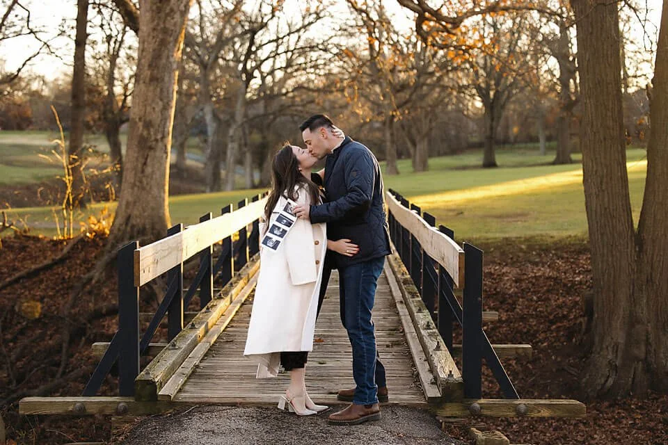 Pregnancy Announcement Photos capturing a sweet kiss between an expecting couple on a rustic bridge surrounded by golden fall foliage