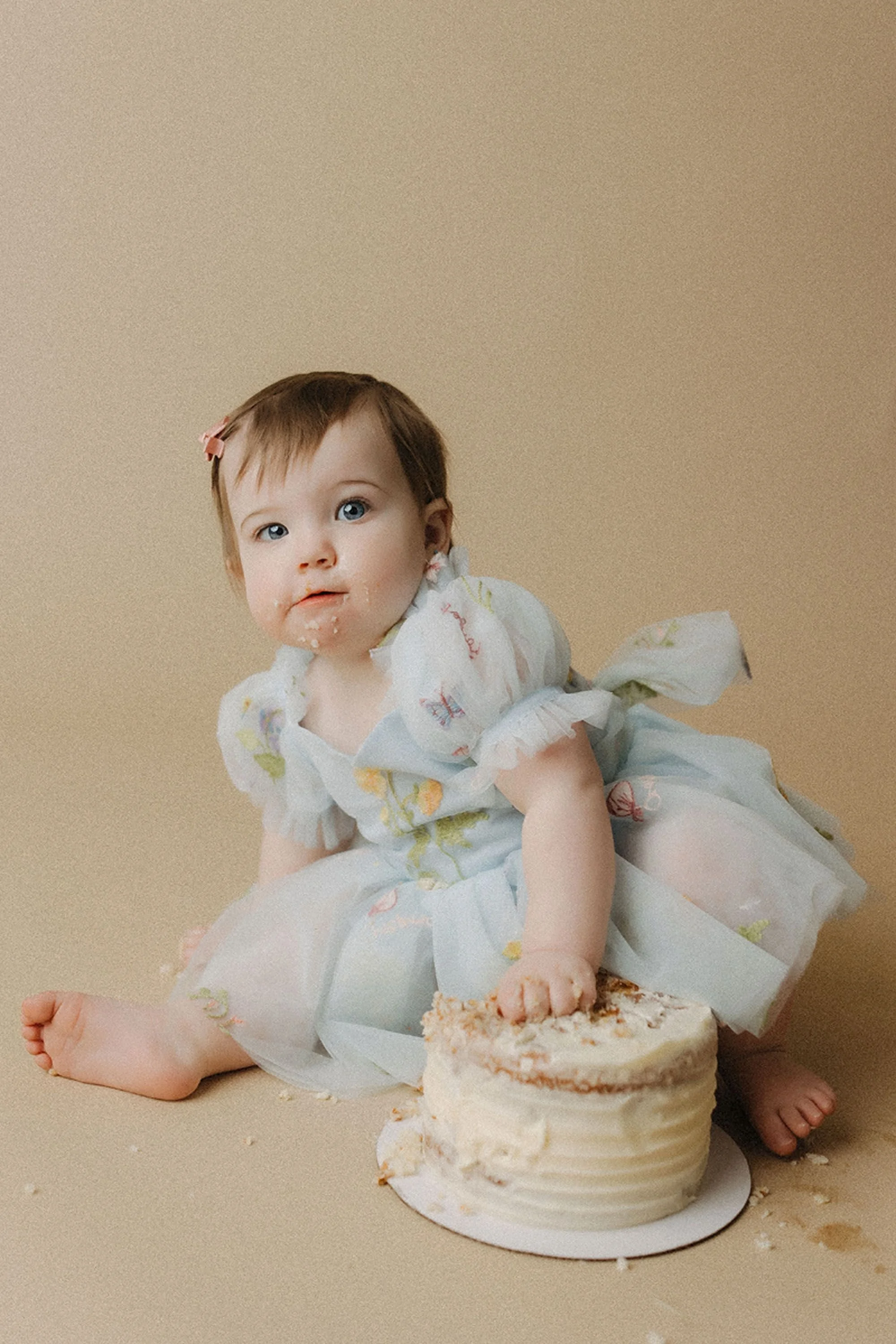 Simple Cake Smash Photos of a baby girl in a pastel blue dress exploring her first birthday cake with frosting on her face, captured in a warm and minimalist studio setting.