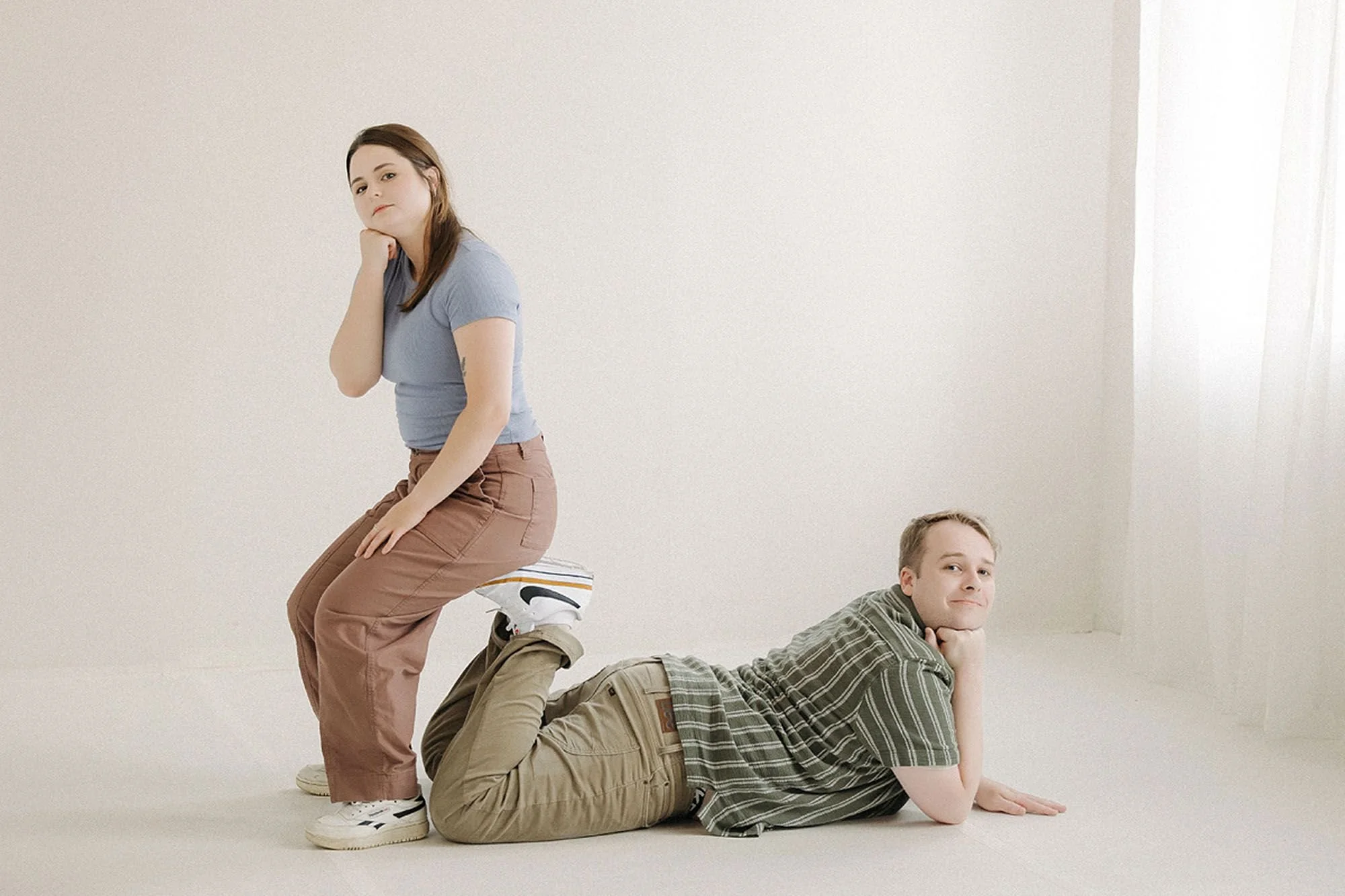 Playful sibling studio portrait a young woman crouches with her chin resting on her hand while a man lies on the floor with his feet propped up in the air, both looking at the camera with relaxed, cheeky expressions against a plain backdrop