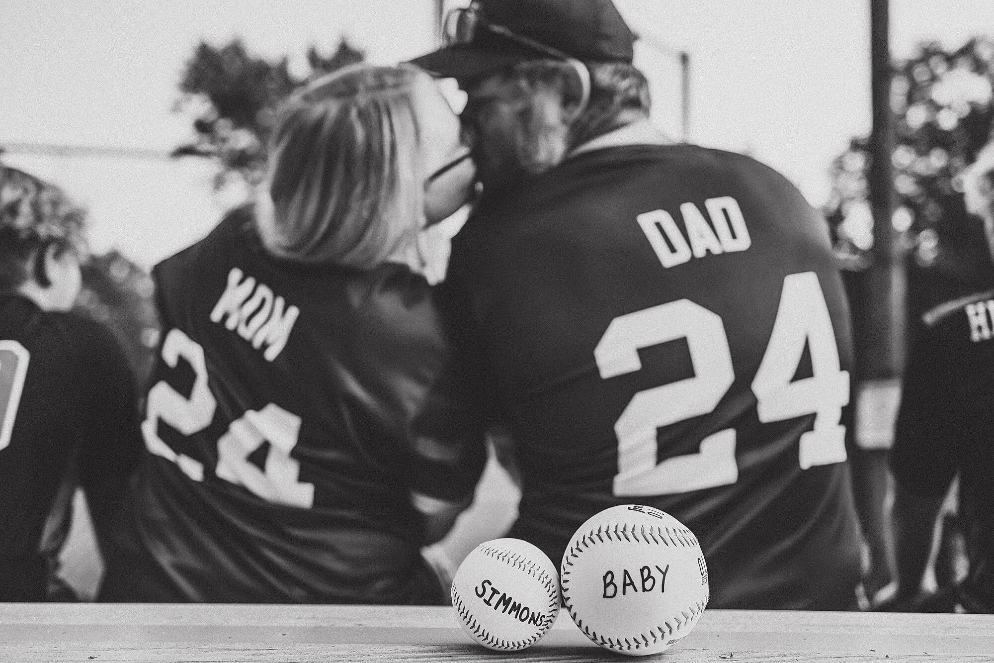 A couple kisses on a bench filled with baseballs, joyfully announcing their pregnancy alongside their older kids