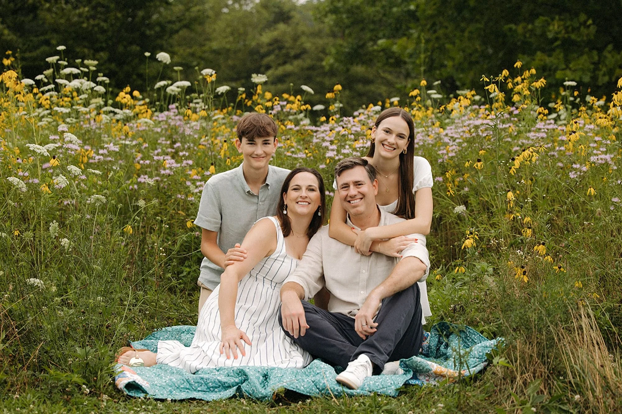 A family of four sitting together on a blanket in a wildflower field during a summer session.