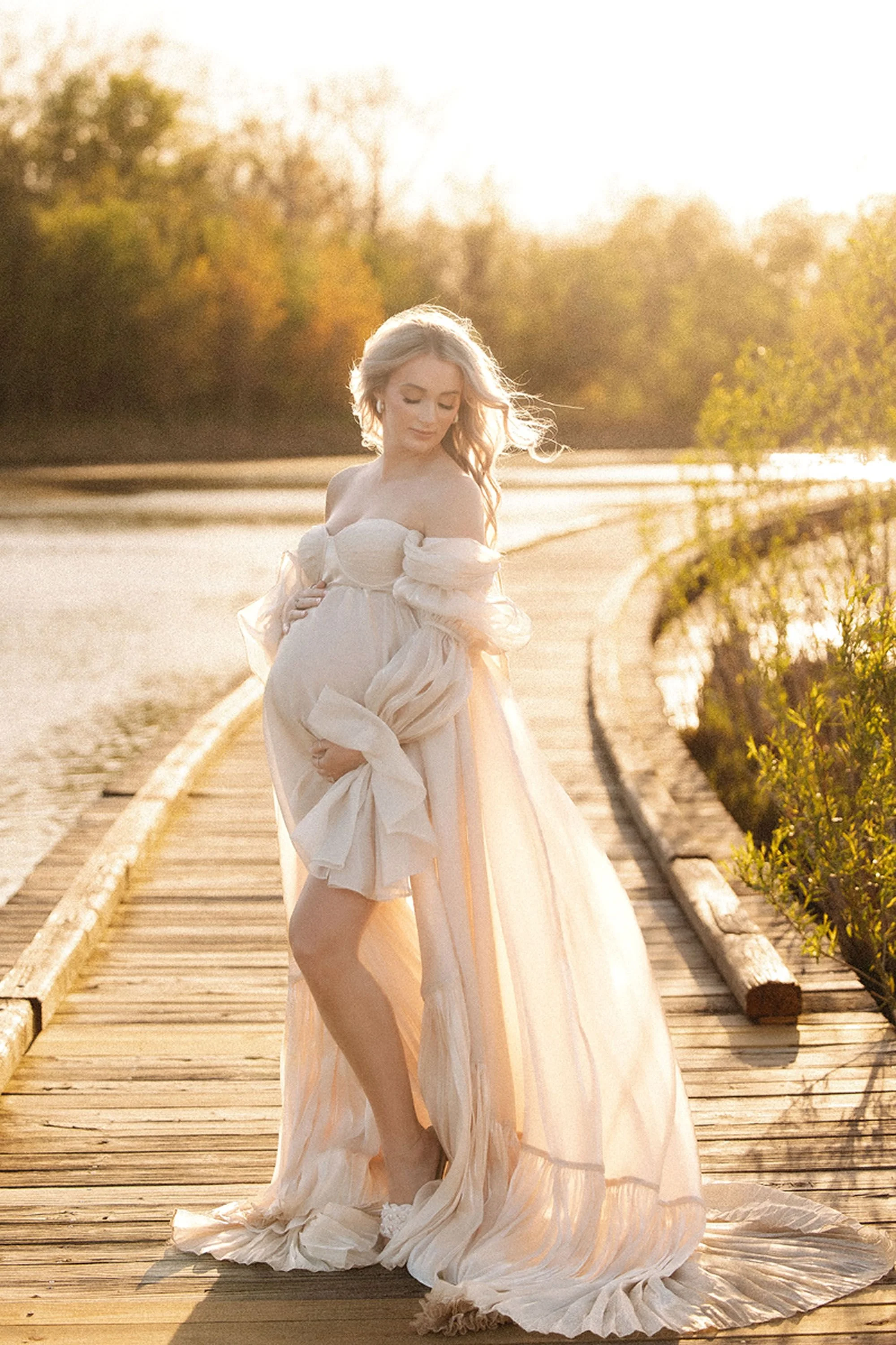 Golden hour maternity photos in Central Indiana featuring a pregnant woman in a flowing ivory gown on a wooden boardwalk by the water, cradling her baby bump with soft autumn trees in the background