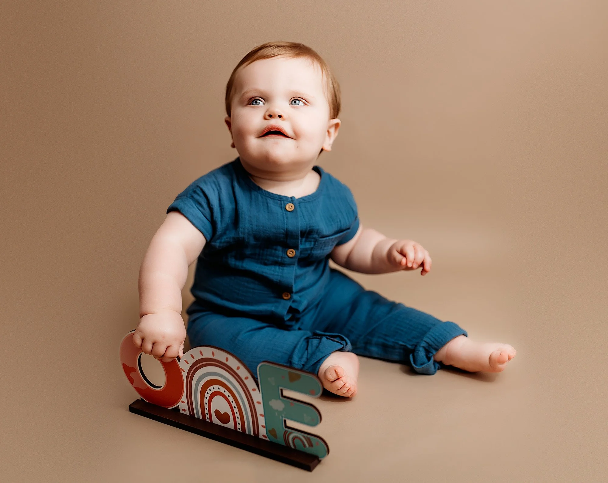 Adorable baby sitting on a beige background holding a decorative wooden word 'LOVE' with rainbow and heart designs, wearing a teal onesie.