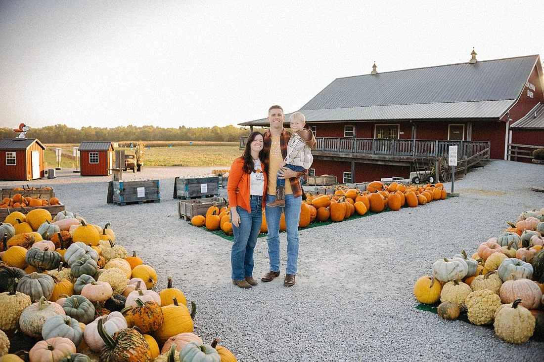 Family of three poses between colorful pumpkin displays outside a rustic red barn during an autumn outing at Dull’s Pumpkin Harvest