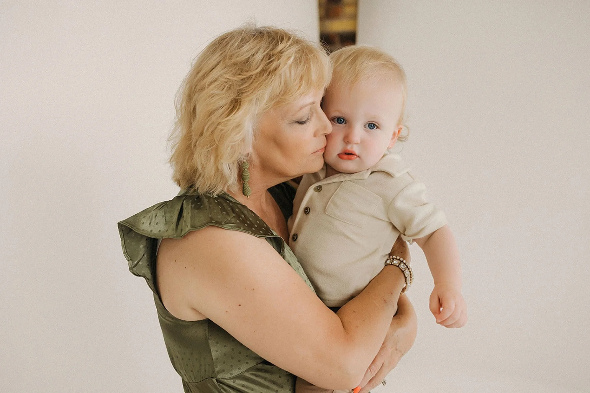 Tender studio portrait of a grandmother in an olive dress cradling her blue-eyed toddler grandson close, gently nuzzling his cheek as he gazes at the camera