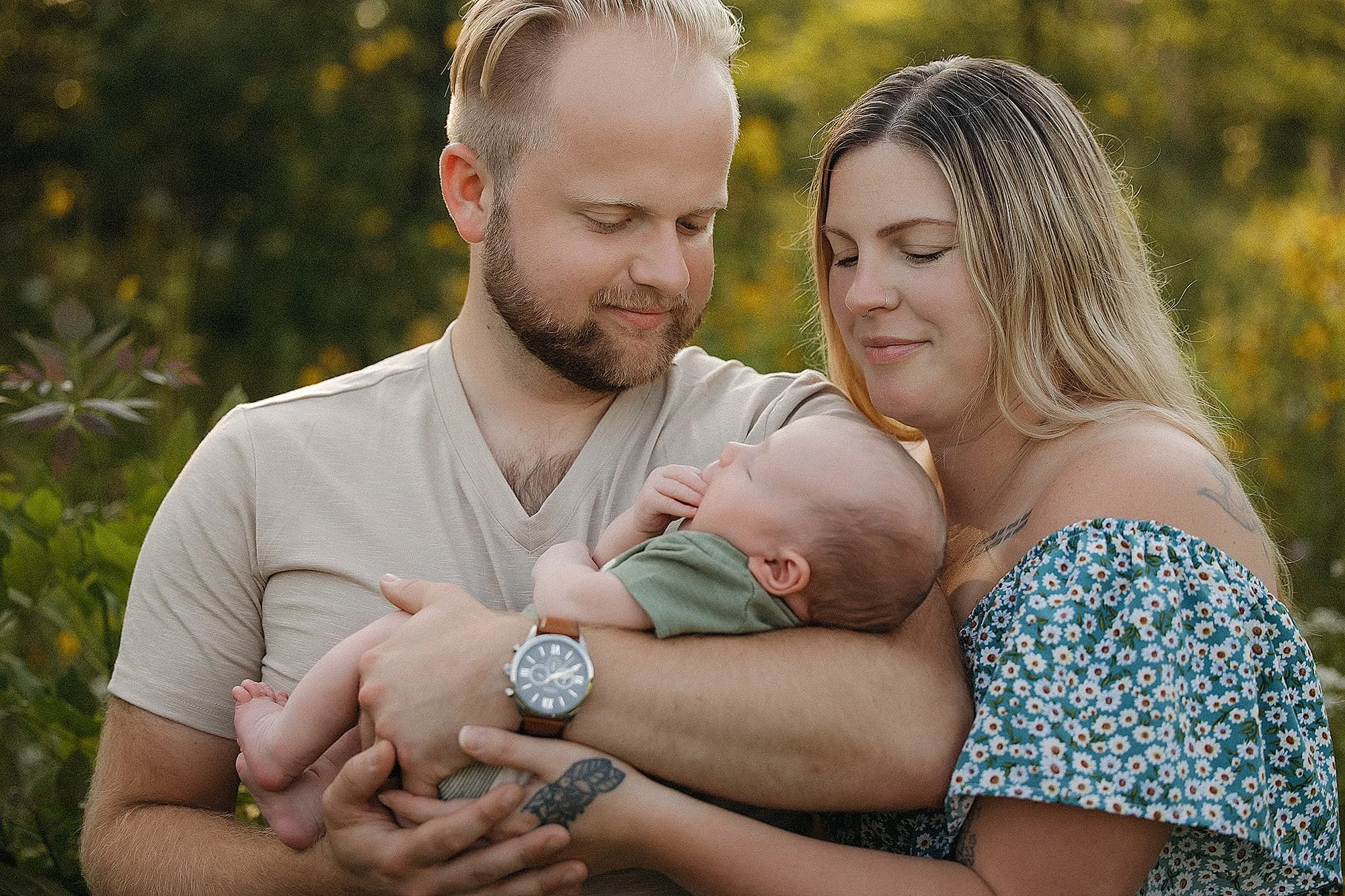 A happy couple holding their newborn baby outdoors during daytime, surrounded by greenery and trees with yellow leaves, close together with gentle smiles.