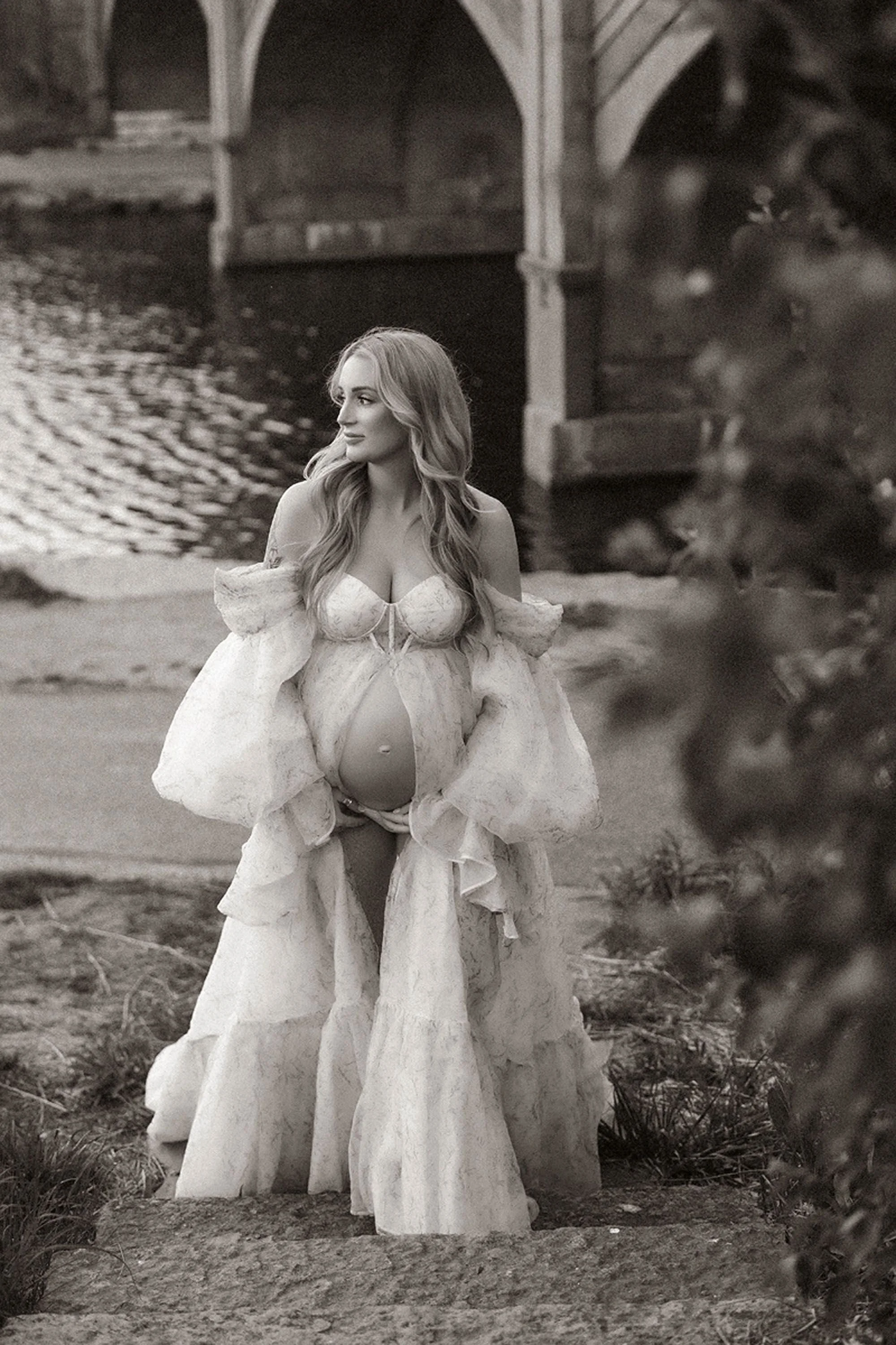 Planning Maternity Photos captured in a timeless black-and-white portrait, featuring an expectant mother in a flowing floral gown standing outdoors and looking peacefully toward the soft natural light
