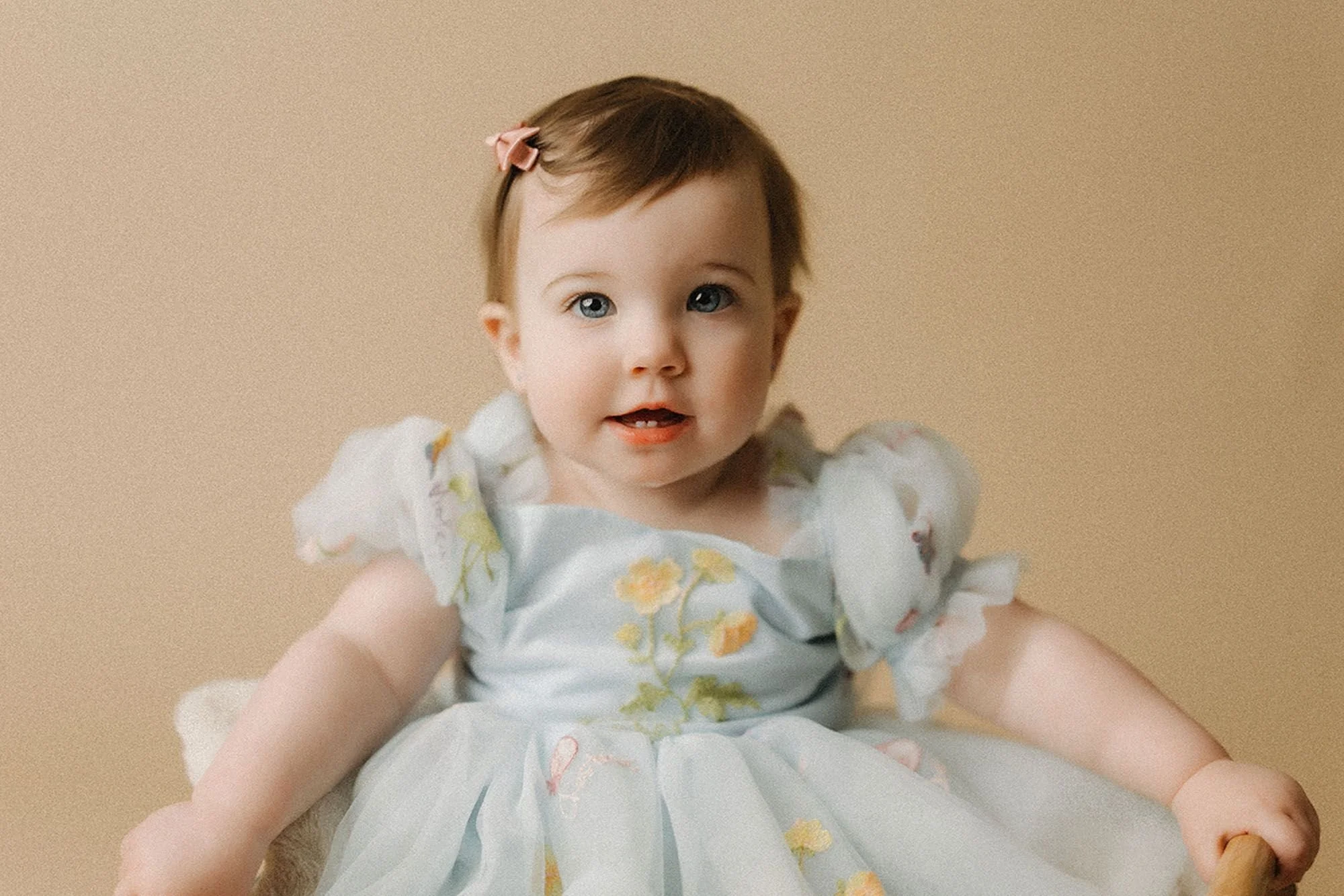 Baby girl in a light blue floral dress with a pink bow, smiling softly at the camera, formal portrait before a simple cake smash photography session