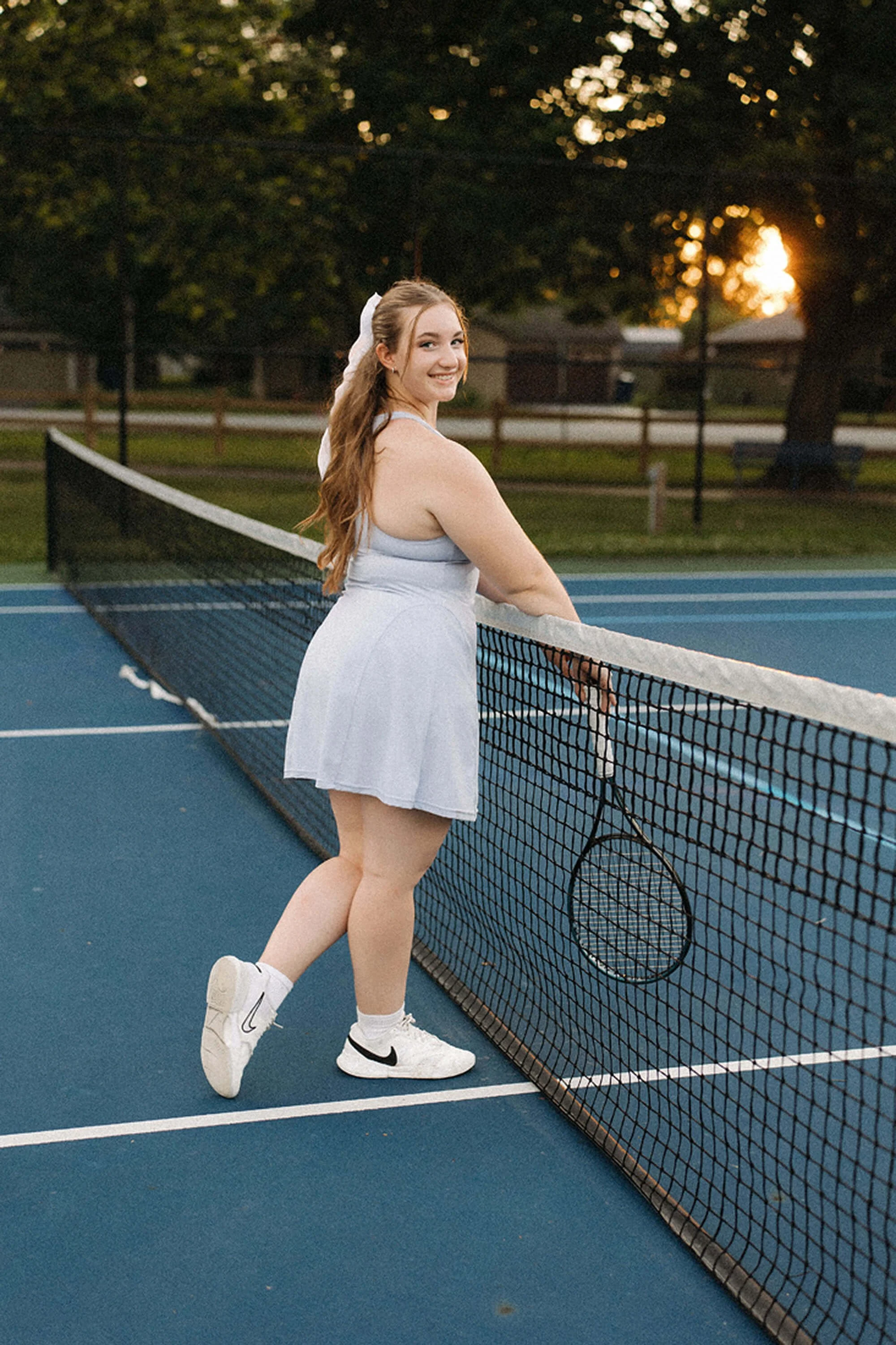 Senior Photos featuring a smiling senior girl posing with her tennis racket by the net during a warm sunset session