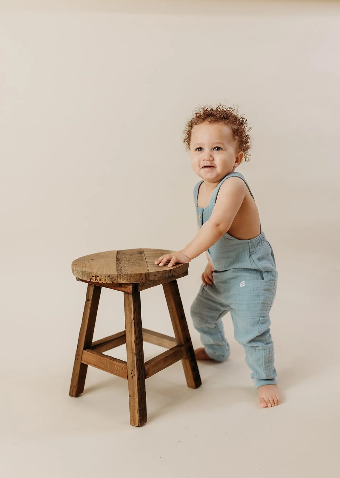 A young toddler with curly hair wearing a light blue sleeveless romper, standing barefoot, holding onto a small wooden stool, with a curious expression on his face against a plain light background.