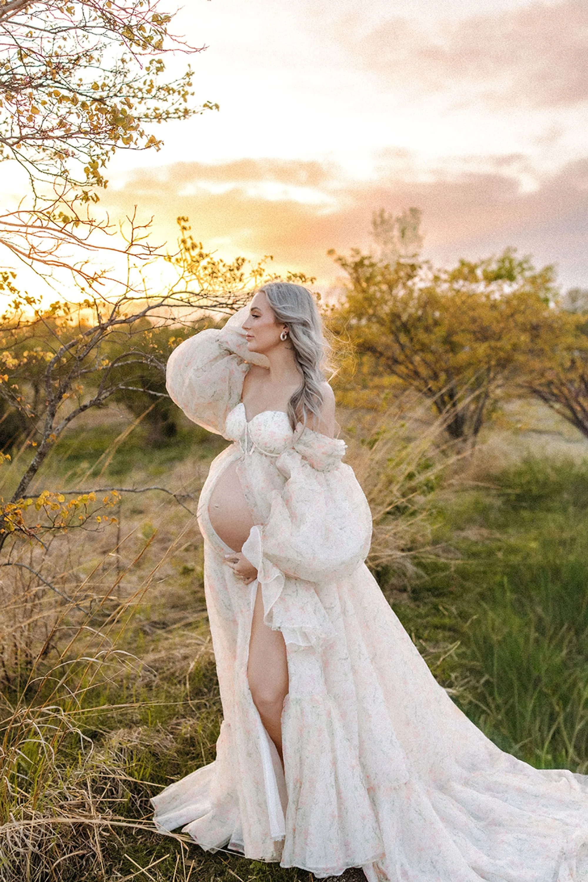 Pregnant woman's side profile against a golden sunset sky, wind blowing her hair and floral gown in an open field during a maternity photo session in Central Indiana