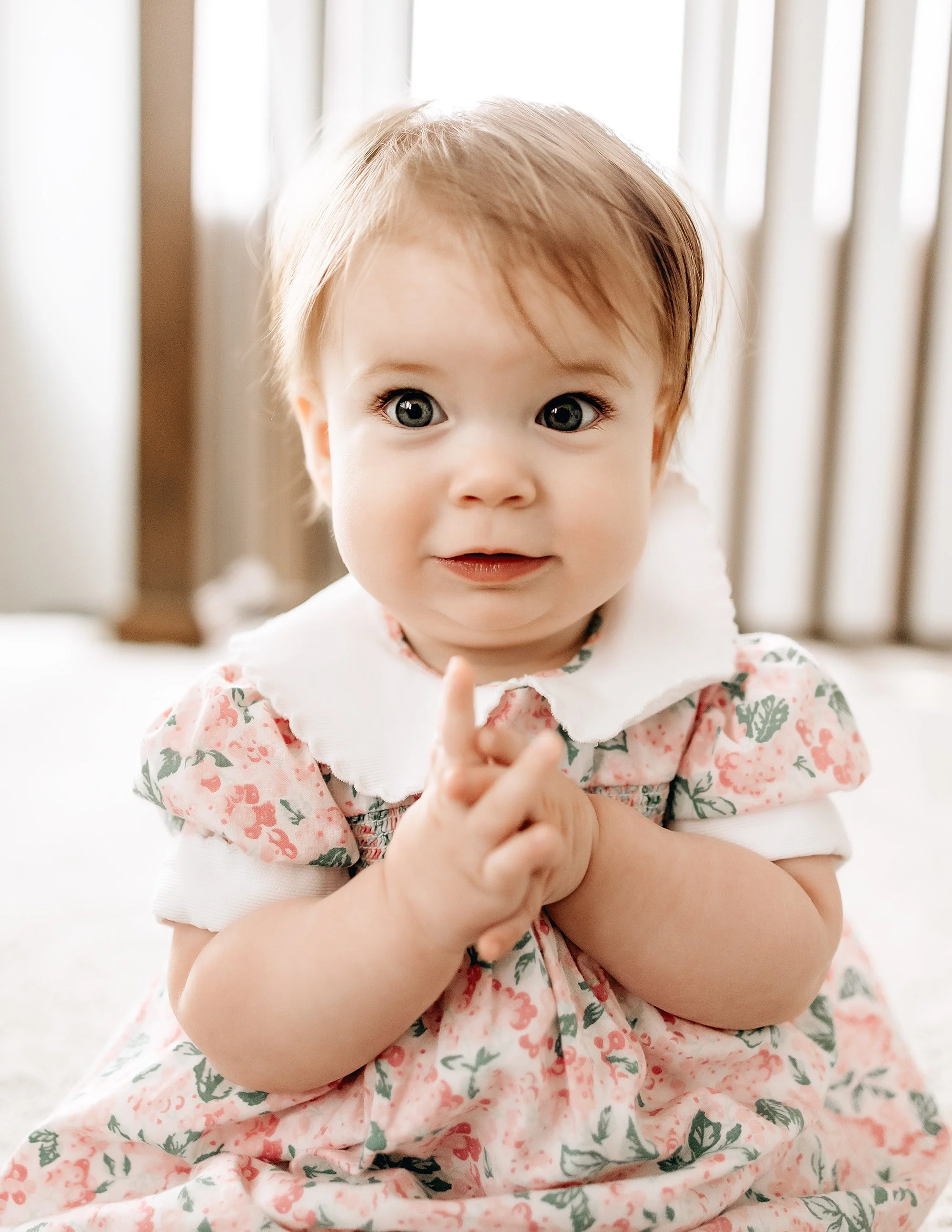 A young girl with big eyes and light brown hair sits on the floor, wearing a floral dress with a white collar, clapping her hands.