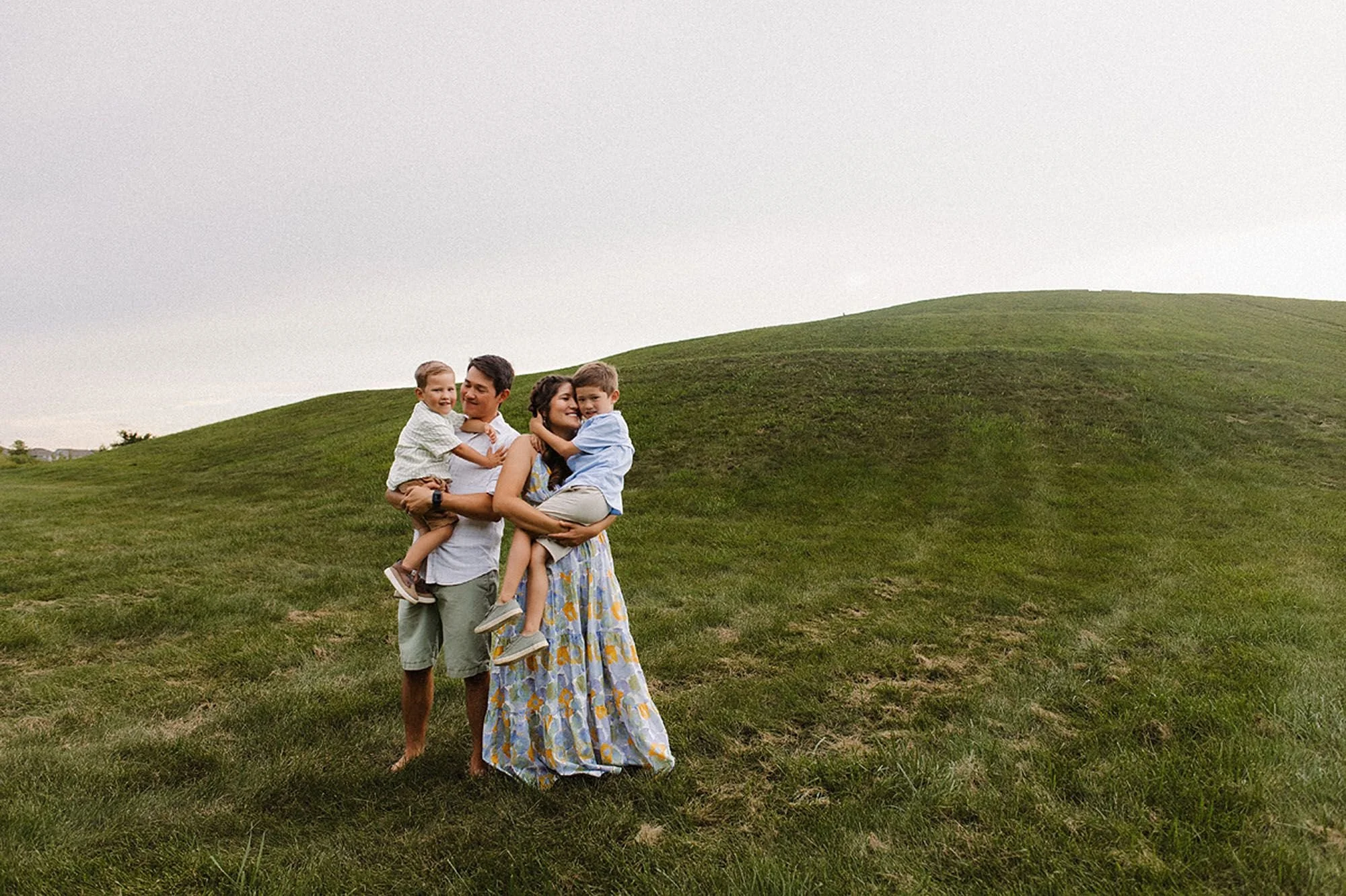 Parents holding their two young sons on a rolling green hill under an overcast sky during a Flat Fork Creek Park family photo session