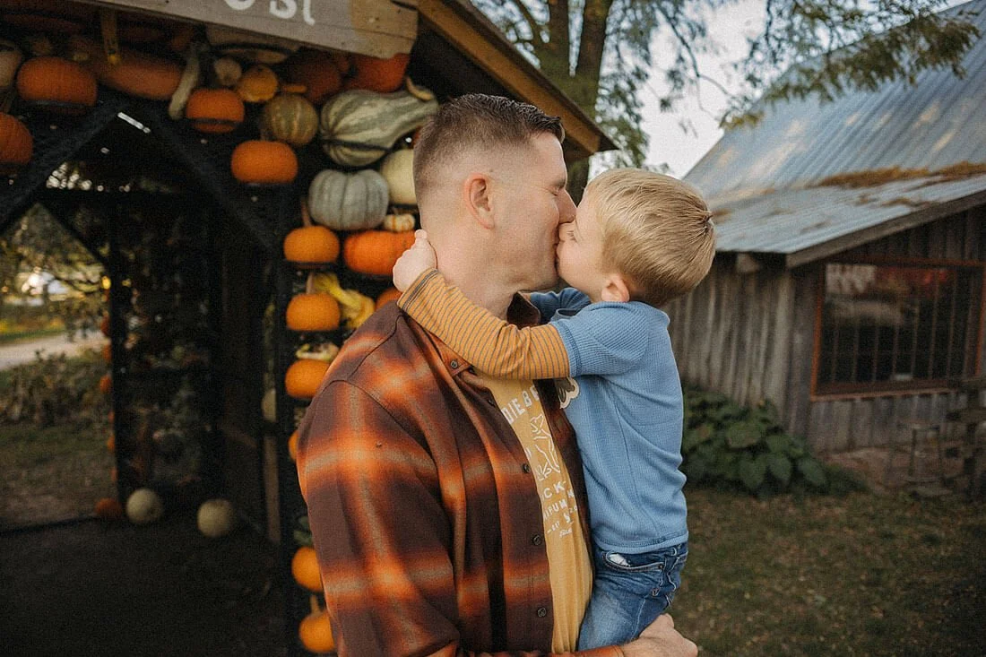 Father holds his young son as they share a kiss at Dull’s Pumpkin Harvest, beside a colorful pumpkin display and rustic wooden building