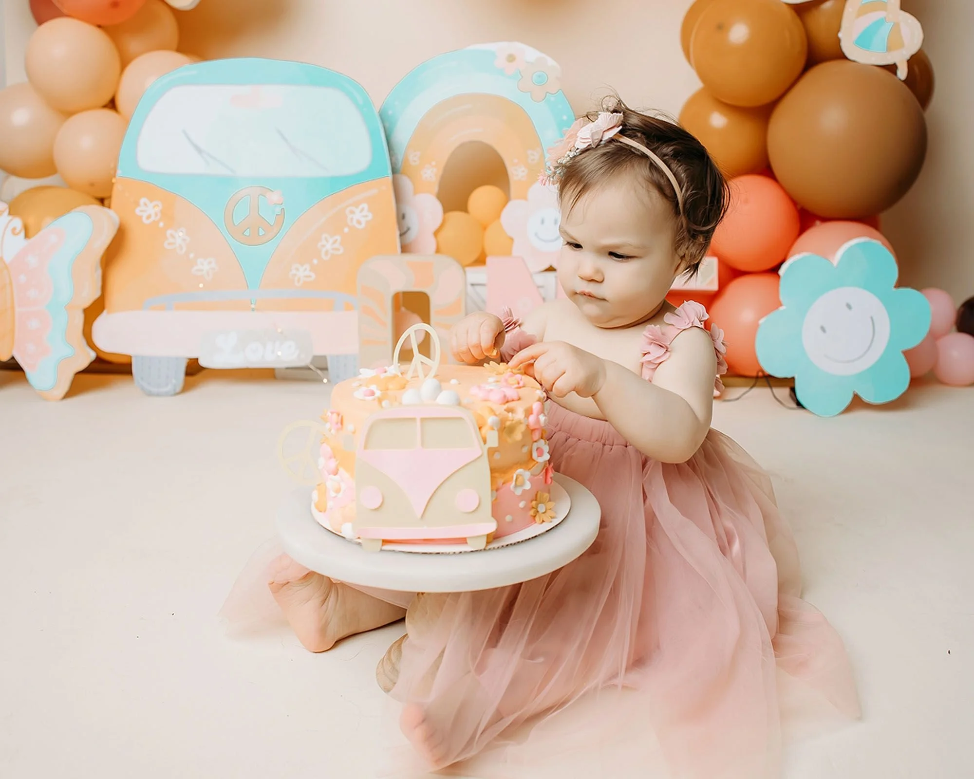 Toddler girl in a blush pink dress enjoying a retro groovy one cake smash session with a VW bus cake and colorful boho balloons in an Indianapolis photography studio