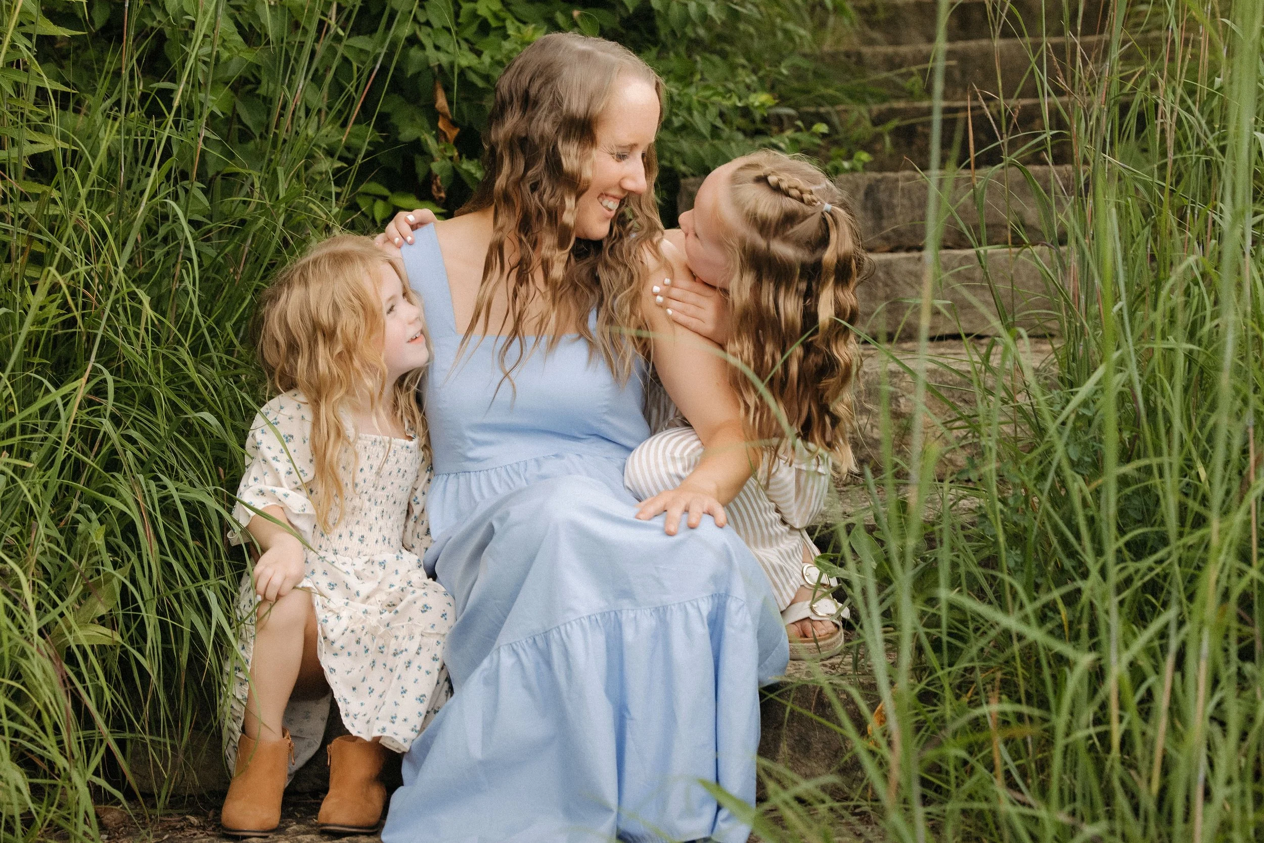 A woman and two girls relax on steps amidst tall grass, creating a warm family atmosphere in this extended family photo