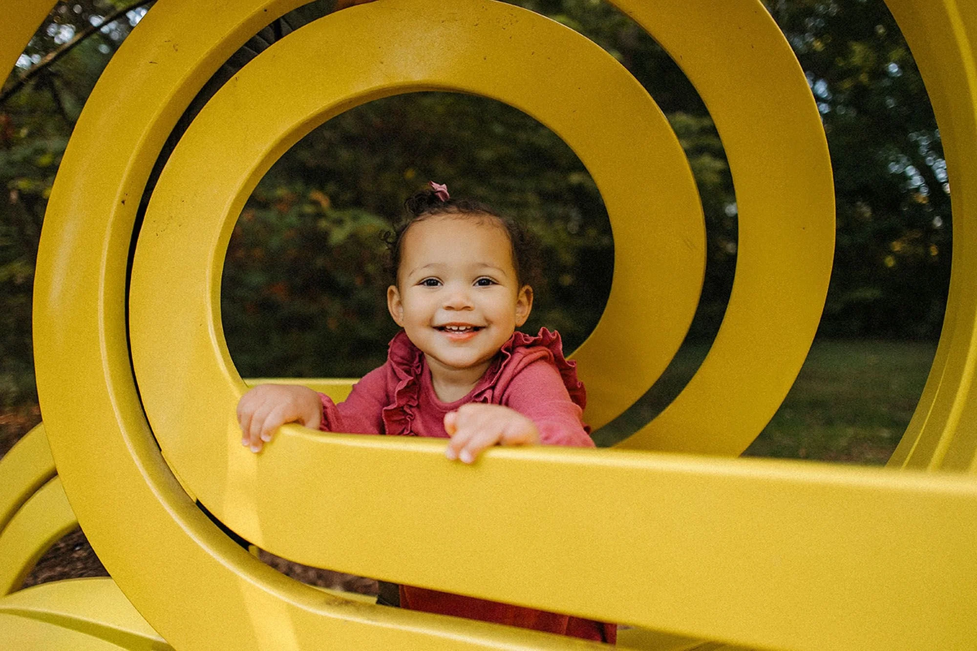 Close-up of a smiling toddler in a pink ruffled top framed by the concentric circles of a yellow spiral sculpture at Virginia B. Fairbanks Art & Nature Park family photos highlighting playful details up close