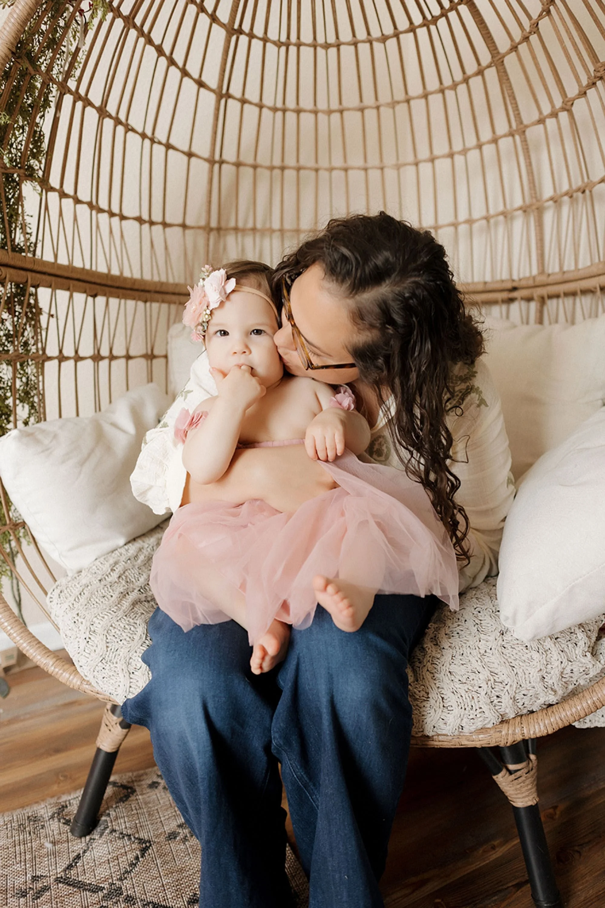 Mother kissing her baby daughter's cheek while sitting together in a woven rattan egg chair, baby wearing a pink tulle dress and floral headband