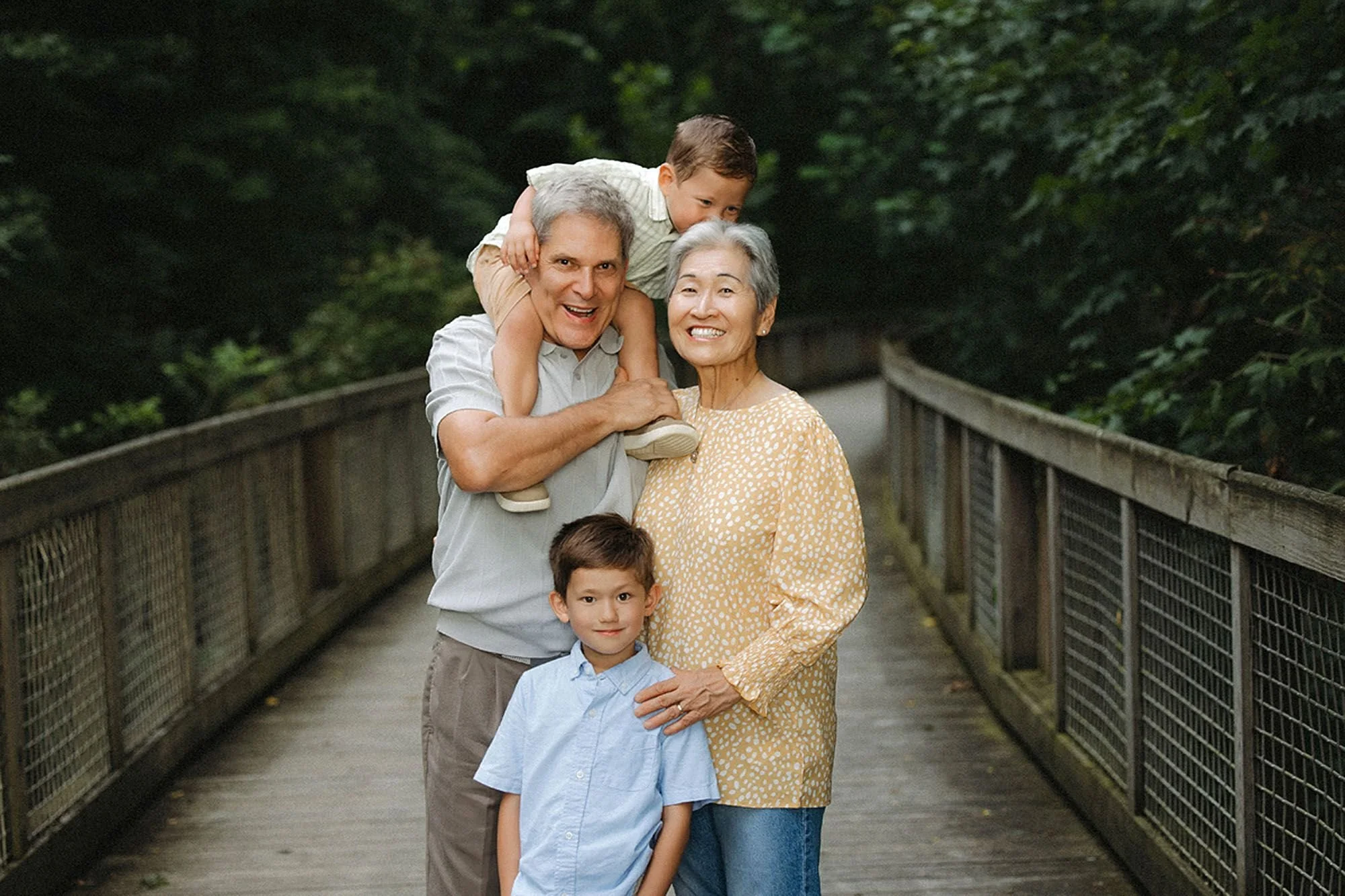 Grandparents smiling with two grandsons on a wooden bridge, one boy on grandfather's shoulders, during a Flat Fork Creek Park family photo session