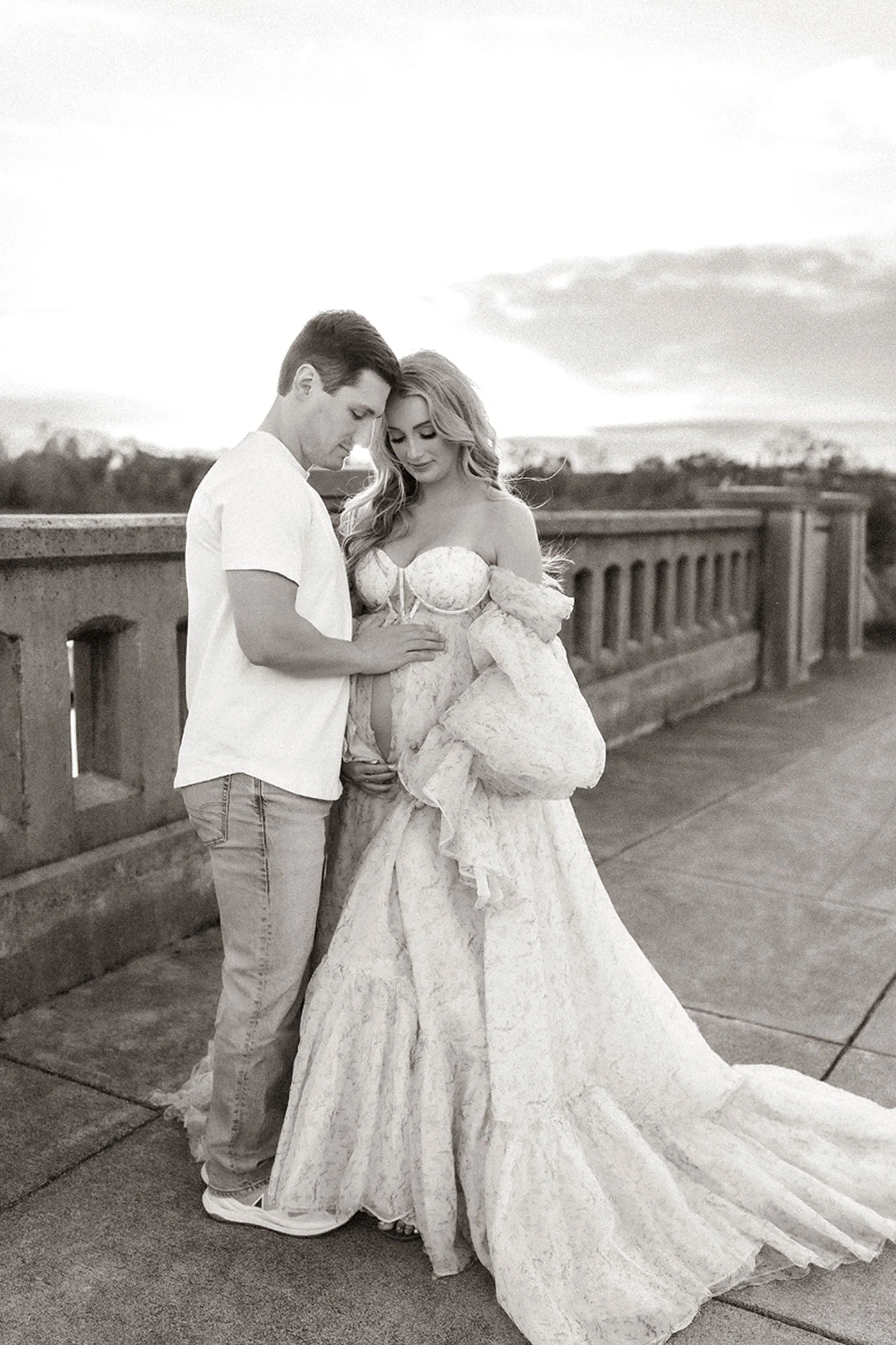Black and white maternity photos in Central Indiana of an expecting couple standing on a stone overlook, husband cradling his wife's baby bump in a lace gown
