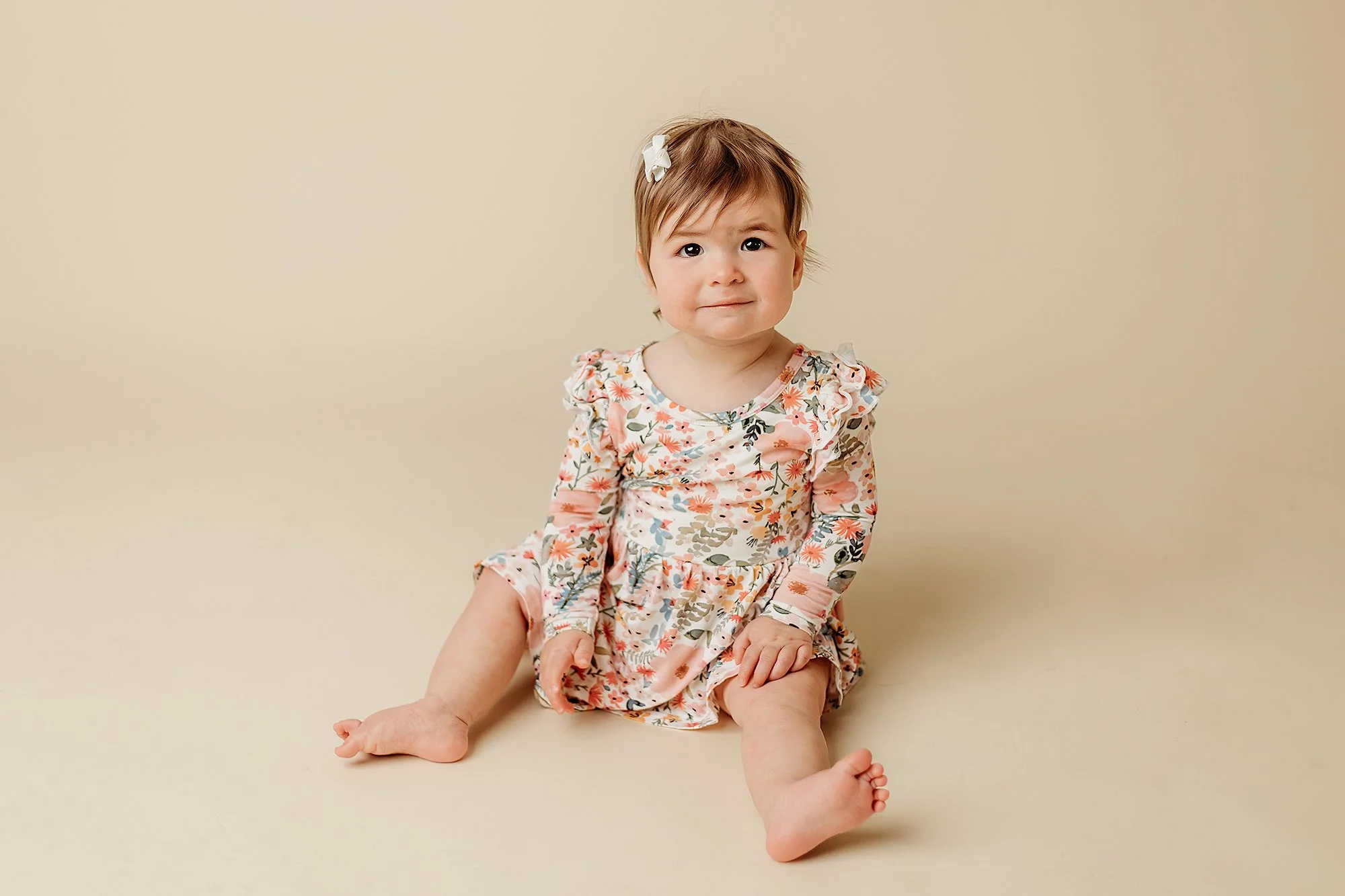Young girl with short brown hair, wearing a floral dress, sitting on the floor against a beige background.