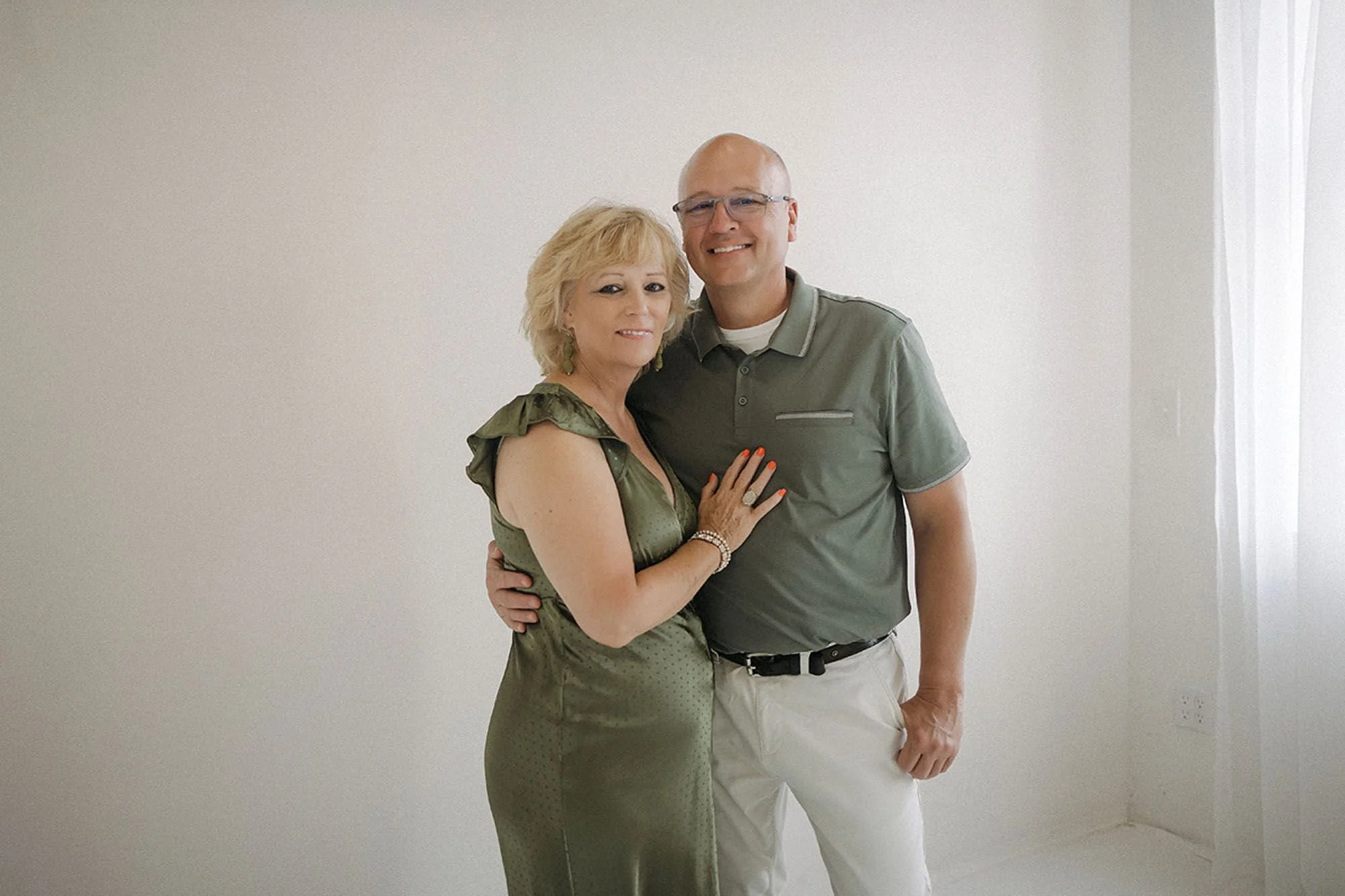 A couple posing closely for a studio portrait, both dressed in matching sage green. She rests her hand on his chest and his arm around her waist as they smile together against a soft neutral backdrop