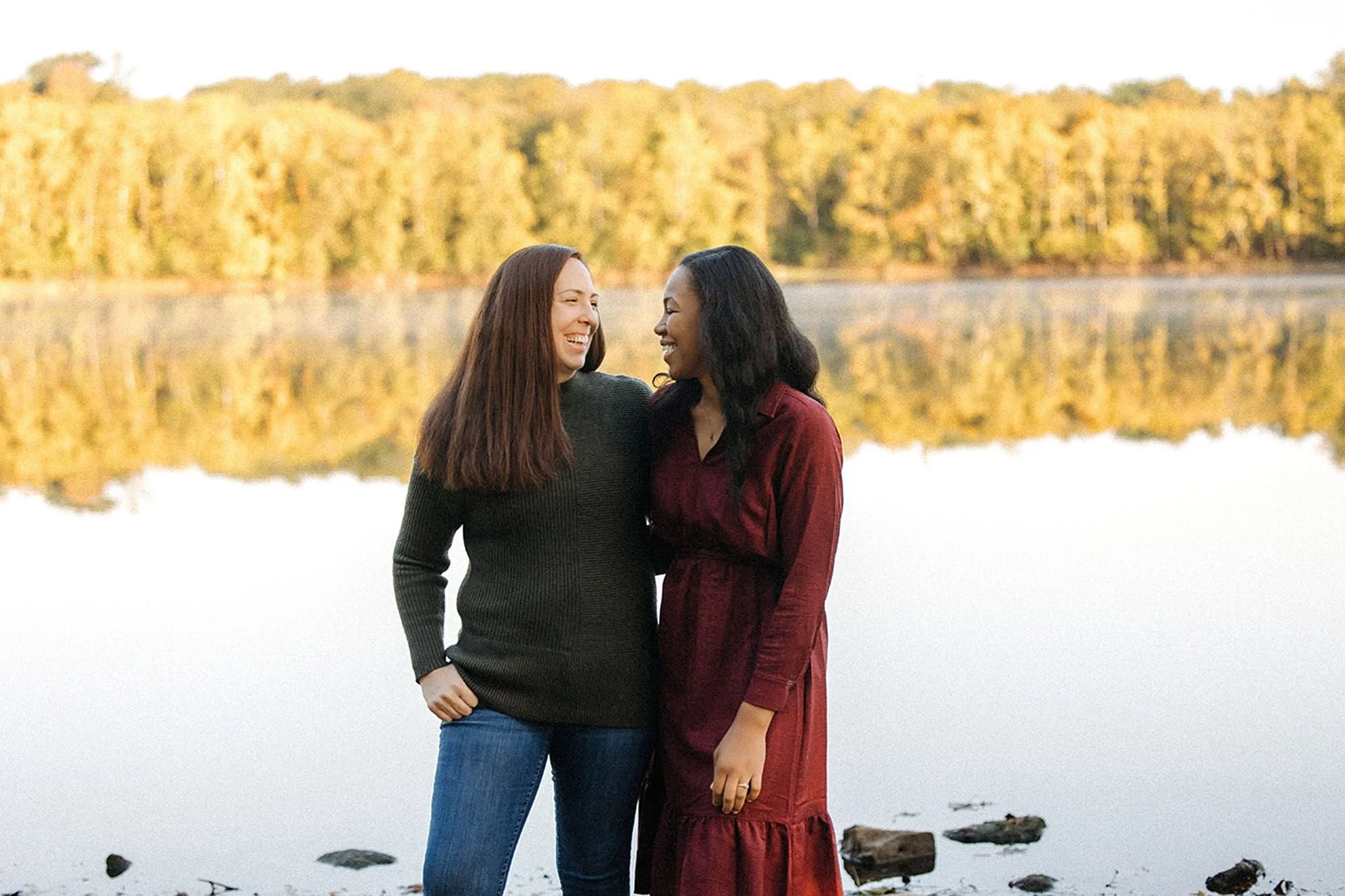 Two women laughing together in front of a still lake with golden autumn trees reflected in the water — Virginia B. Fairbanks Art & Nature Park family photos capturing a warm, joyful moment