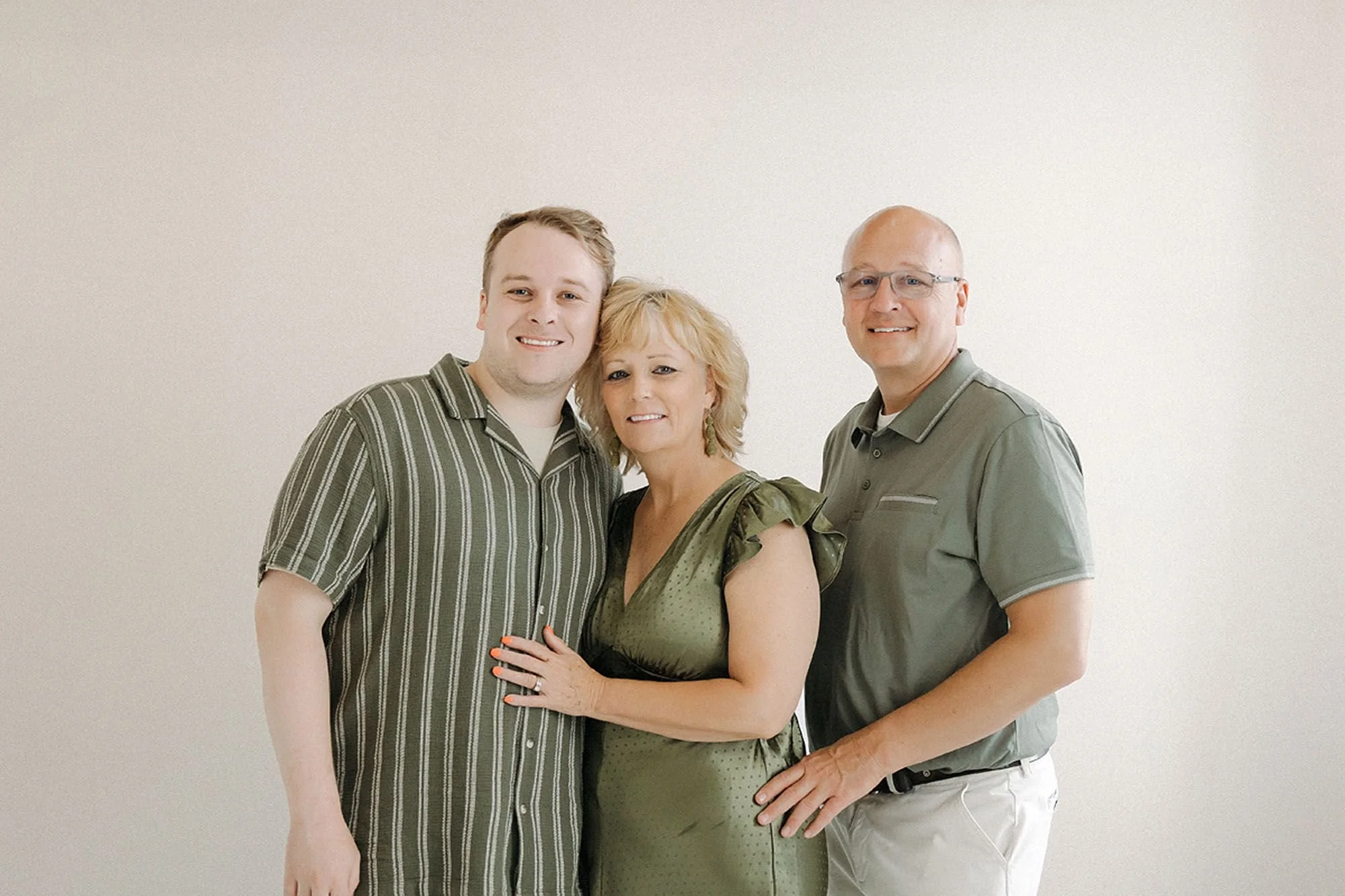 A mother in an olive green dress stands smiling between her husband and adult son, both dressed in coordinating shades of green, with warm affectionate poses against a soft neutral backdrop