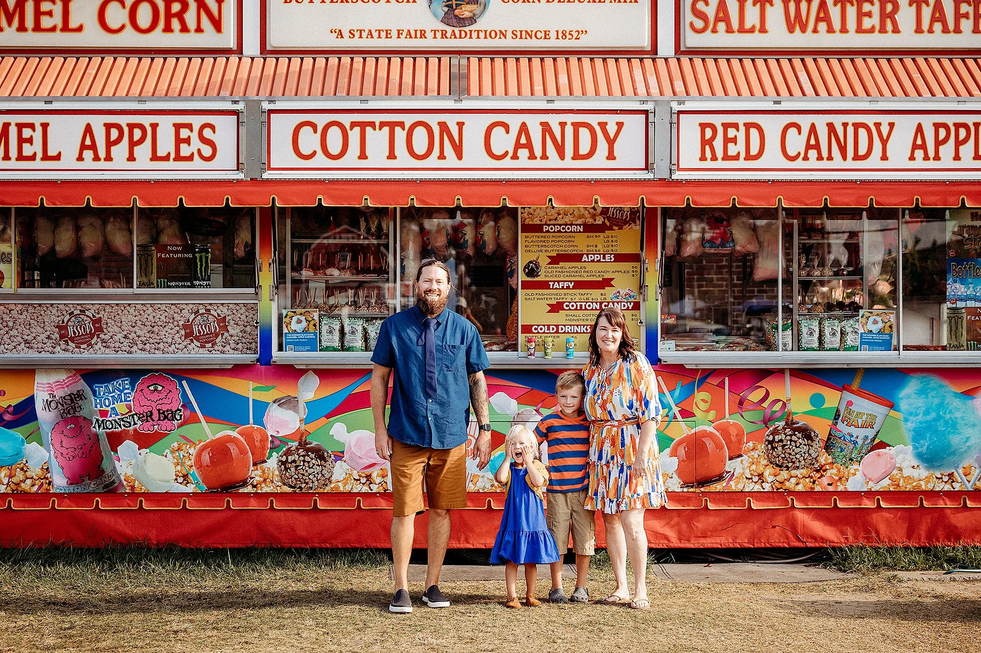 Family posing in front of a colorful cotton candy stand at a county fair, showcasing fun family photo session ideas
