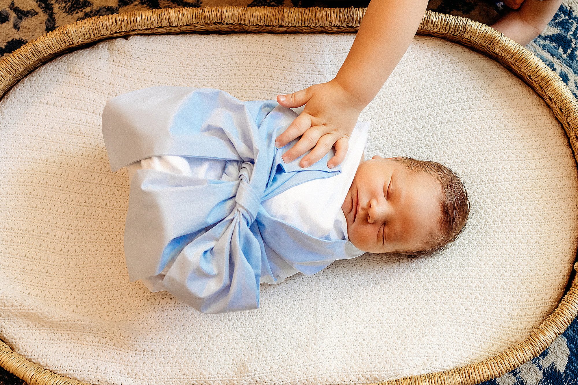Newborn baby sleeping on a cream-colored textured surface inside a woven basket, wearing a white outfit with light blue trim and a bow, with an adult hand gently touching the baby's chest.