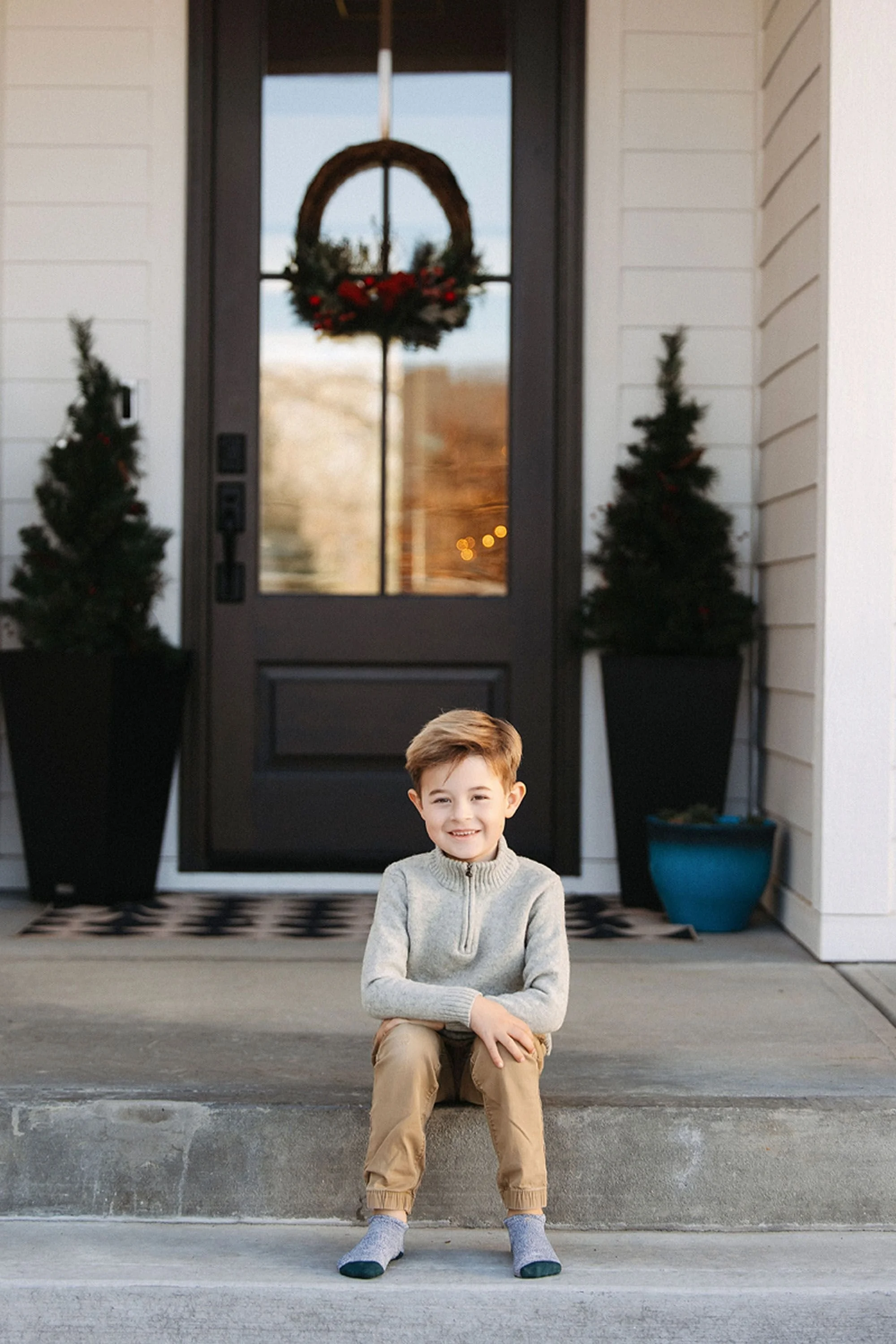 A young boy is smiling on the front steps of his home, surrounded by Christmas trees and festive décor, capturing the warmth of at-home Christmas pictures in Chatham Hills
