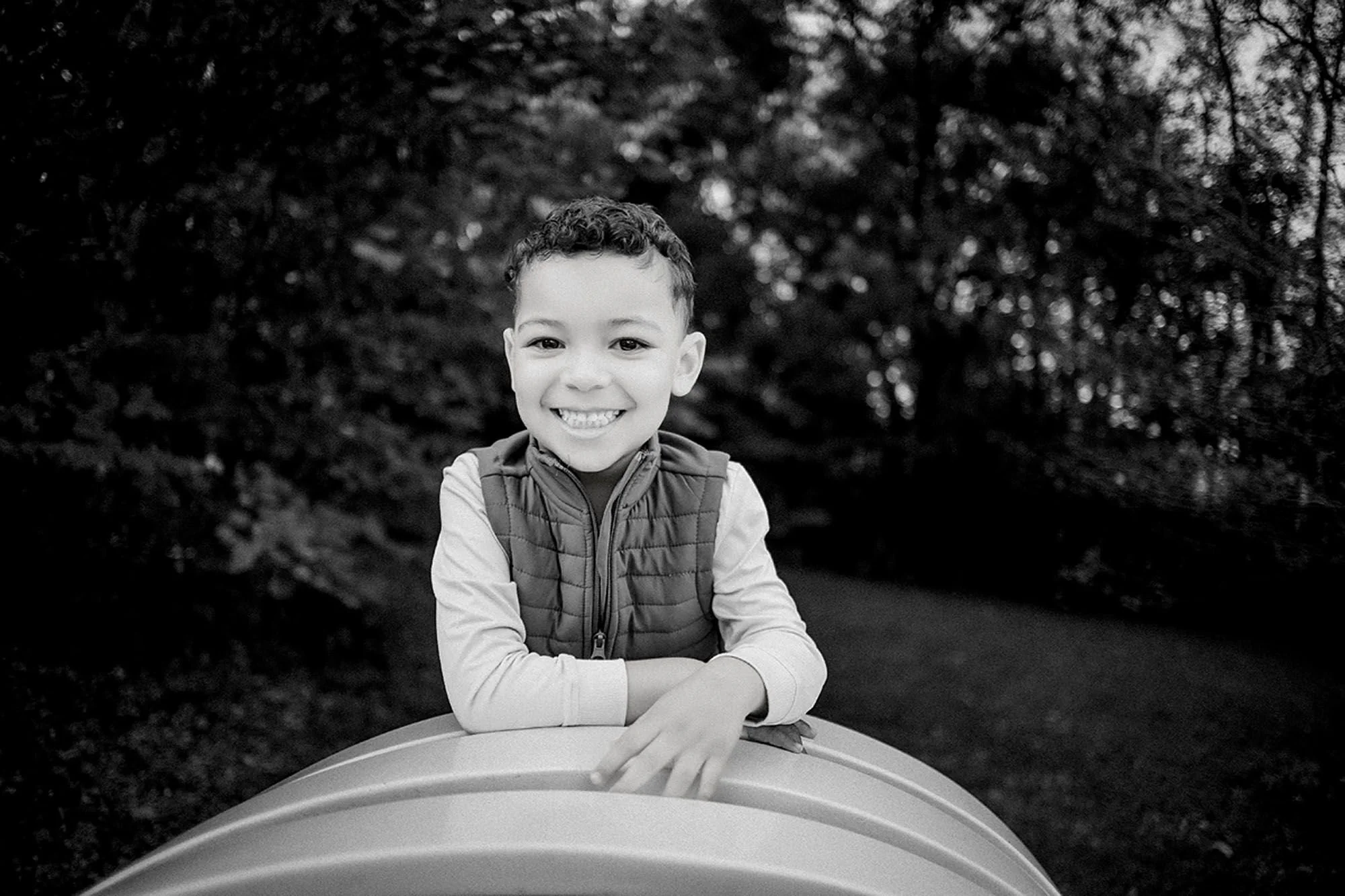 Black-and-white portrait of a smiling young boy in a puffer vest resting his arms on the yellow sculpture bench at Virginia B. Fairbanks Art & Nature Park, family photos featuring a sweet solo kid portrait