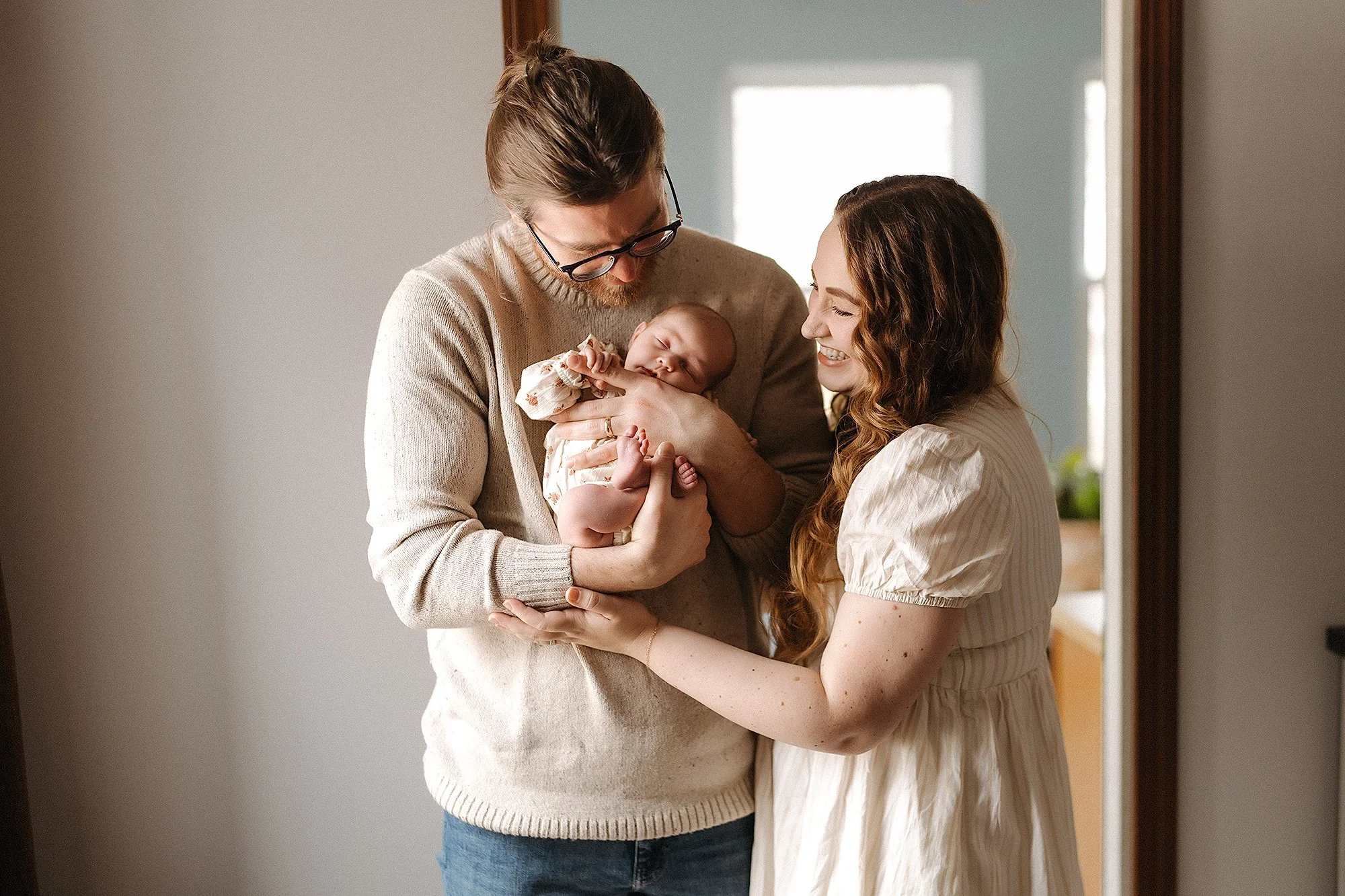 New parents holding their sleeping newborn in a cozy lifestyle home session, wearing soft neutral outfits that showcase what to wear for family photos with timeless elegance, warmth, and natural connection
