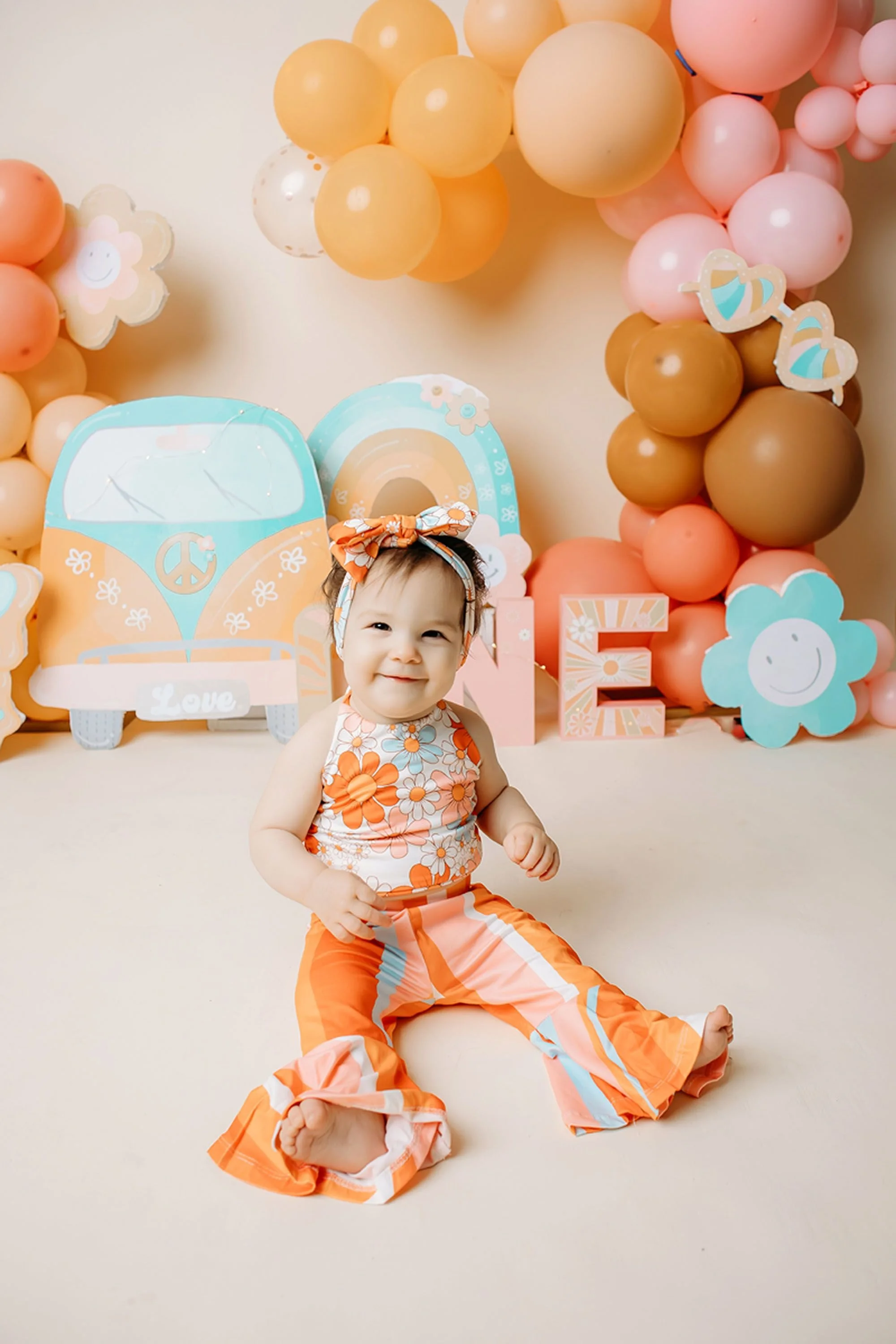 A smiling baby girl sits in a colorful groovy one-themed setup surrounded by orange and pink balloons, retro rainbow decor, and playful floral accents.