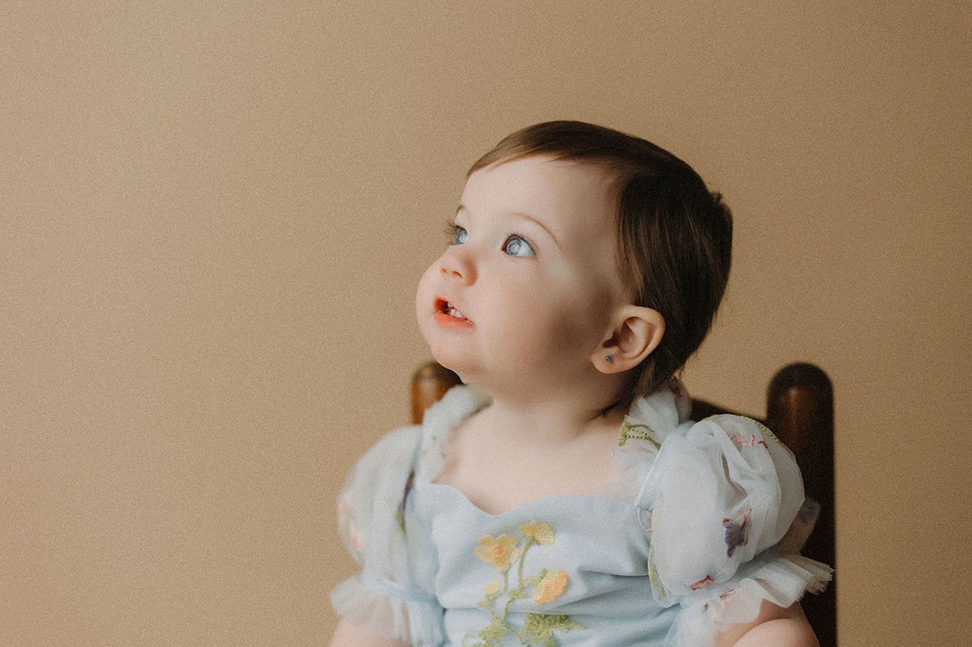 Baby girl in a floral blue dress looking up curiously with wide blue eyes, portrait captured during a simple cake smash photography session