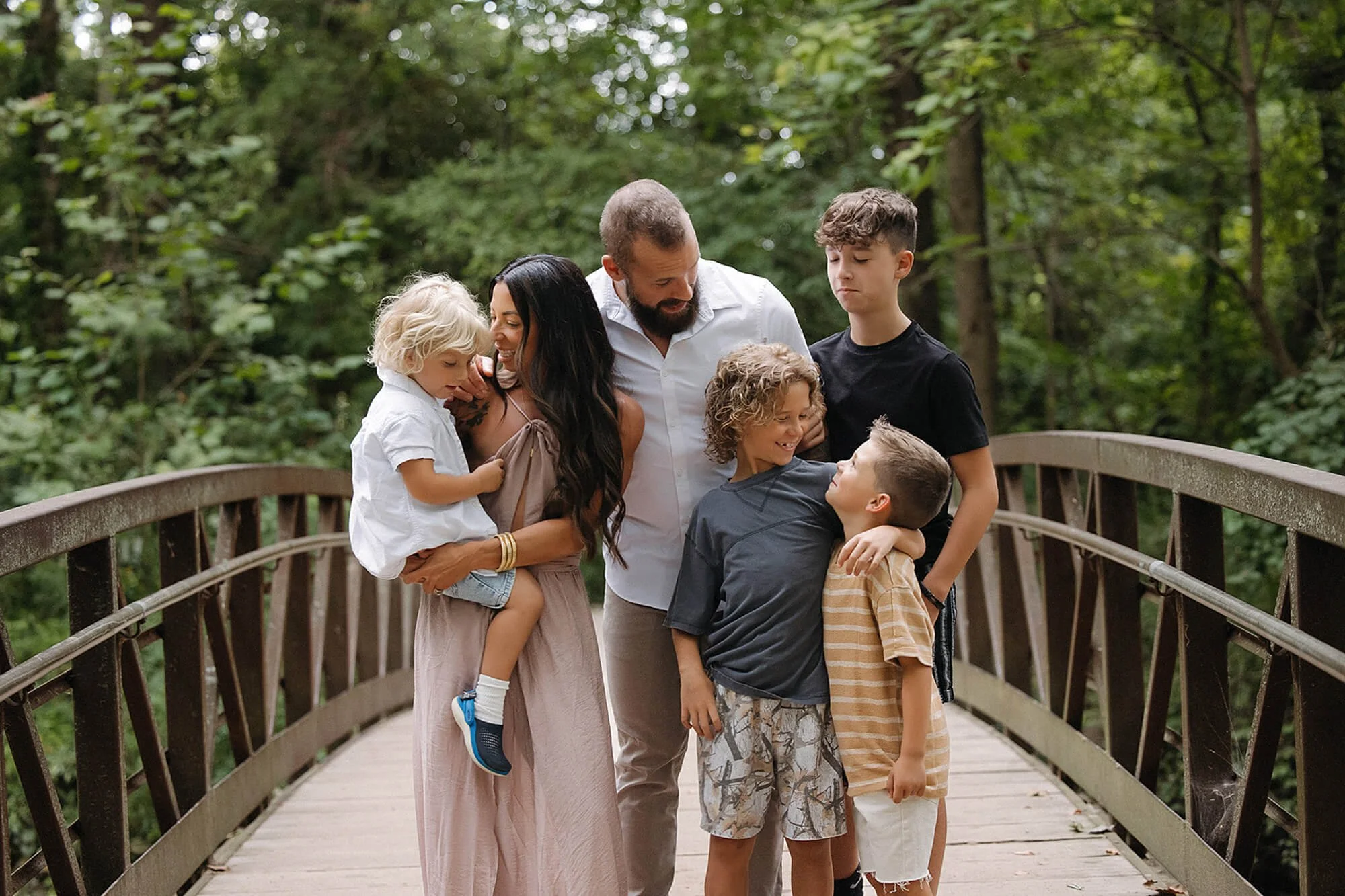 A family stands on a bridge in a wooded area, enjoying a photo session at Flowing Well Park in Carmel, Indiana