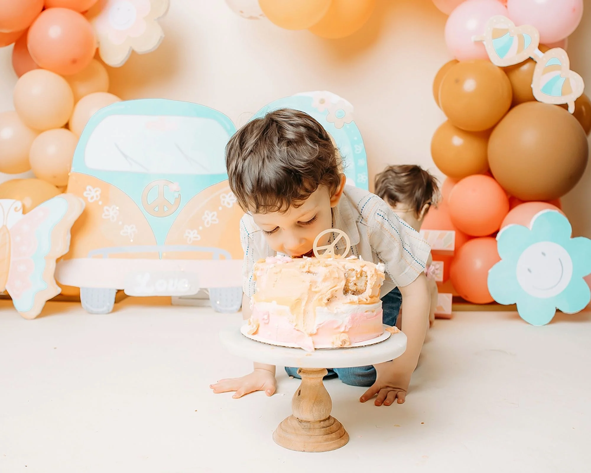 Toddler boy leaning in to bite a pastel pink and cream birthday cake with a peace sign topper, set on a wooden cake stand in front of a retro VW van backdrop and balloon garland