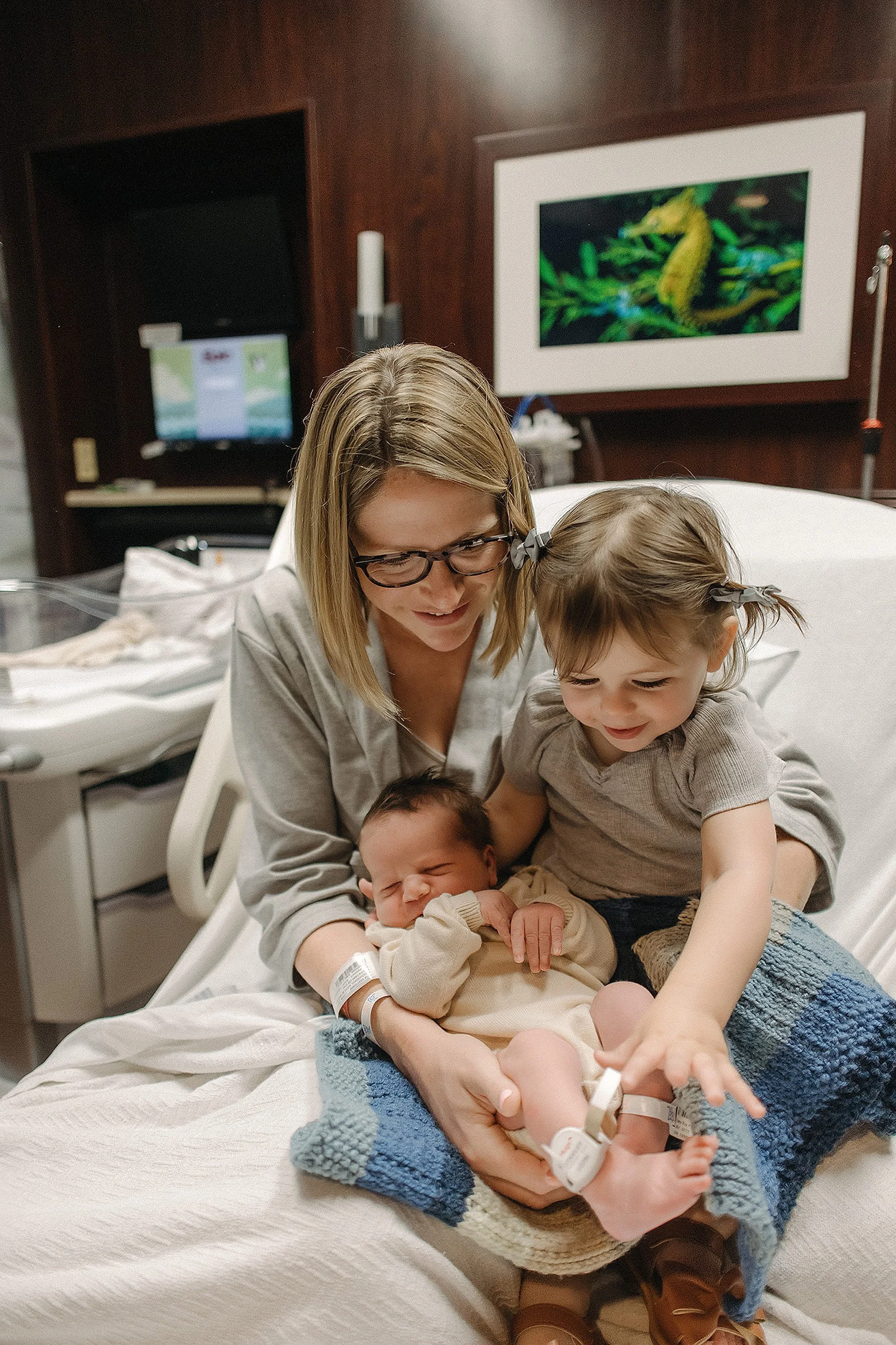 A woman and two young children, a toddler girl and a newborn baby, sitting on a hospital bed in a hospital room, with the woman holding the newborn, and the girl reaching towards the baby's foot.