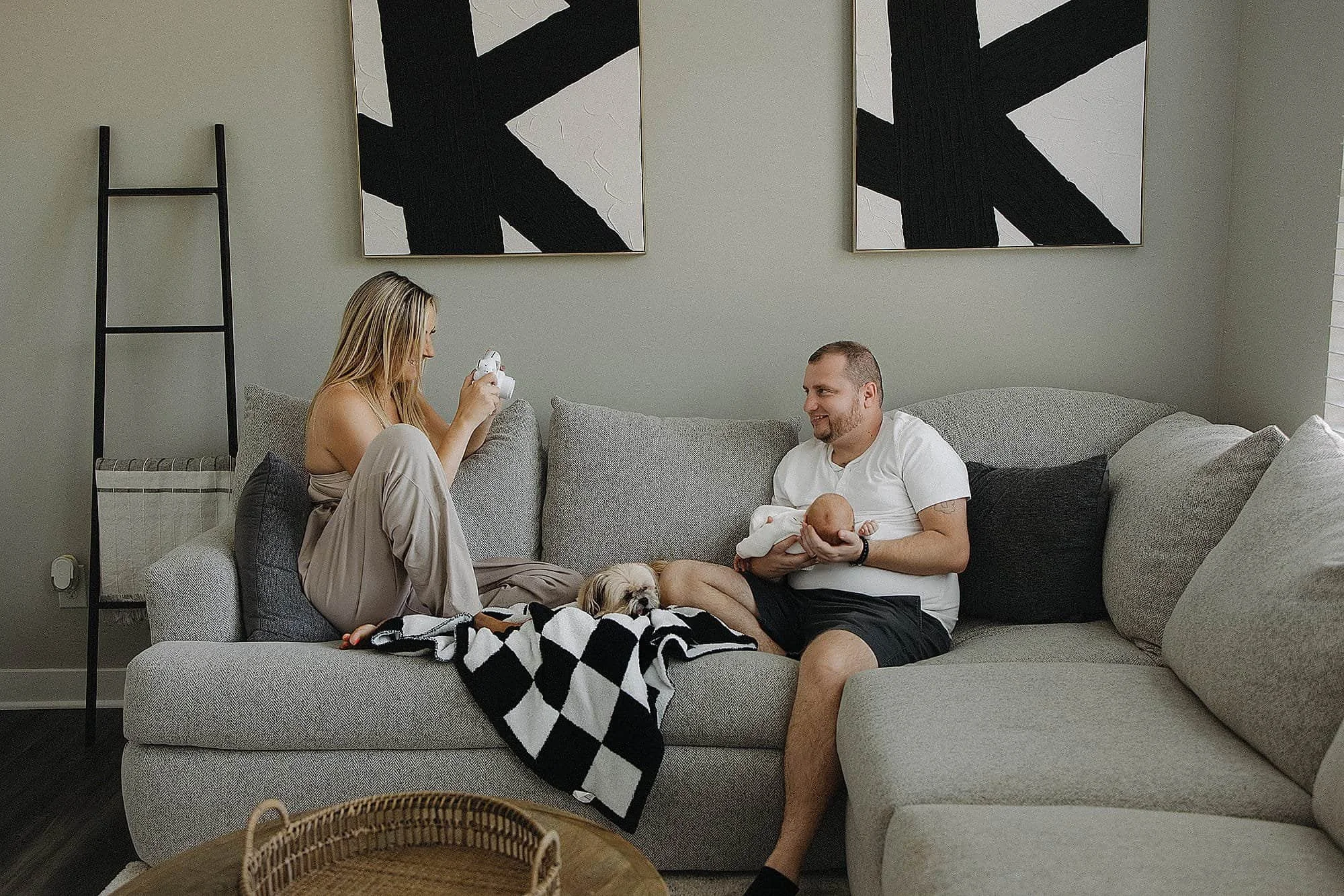 A smiling man holding a newborn baby in a living room with a woman sitting on a couch, a small dog, and modern black and white artwork on the wall.
