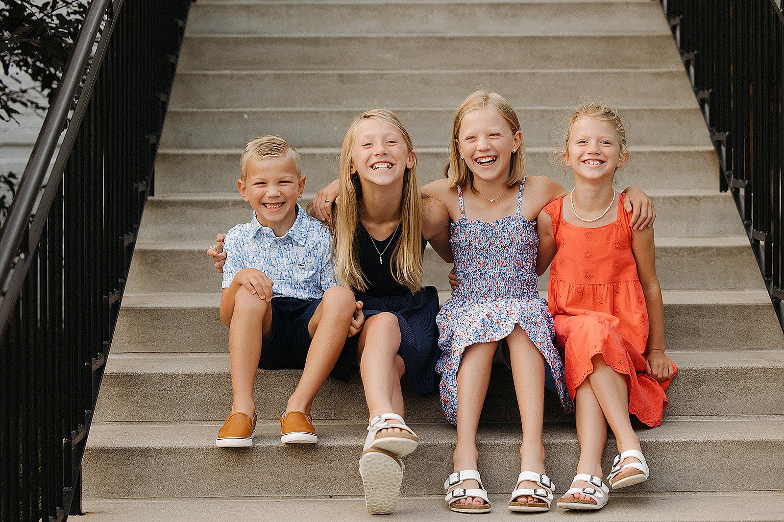 Four siblings sitting together on outdoor steps, smiling and laughing in matching summer outfits.