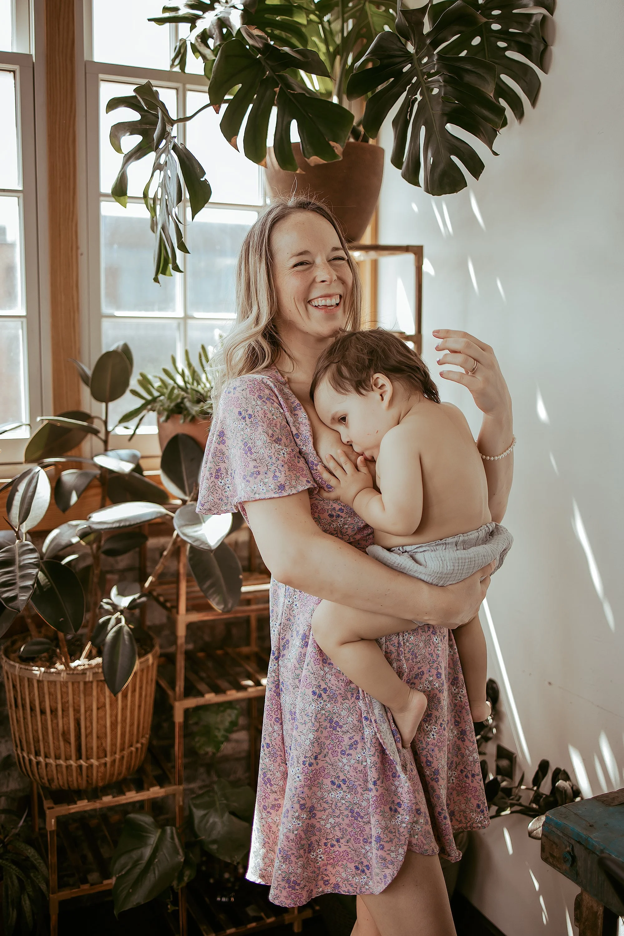 A woman with blonde hair, wearing a floral dress, holding a young boy in her arms inside a room with large windows and several potted plants, smiling happily.