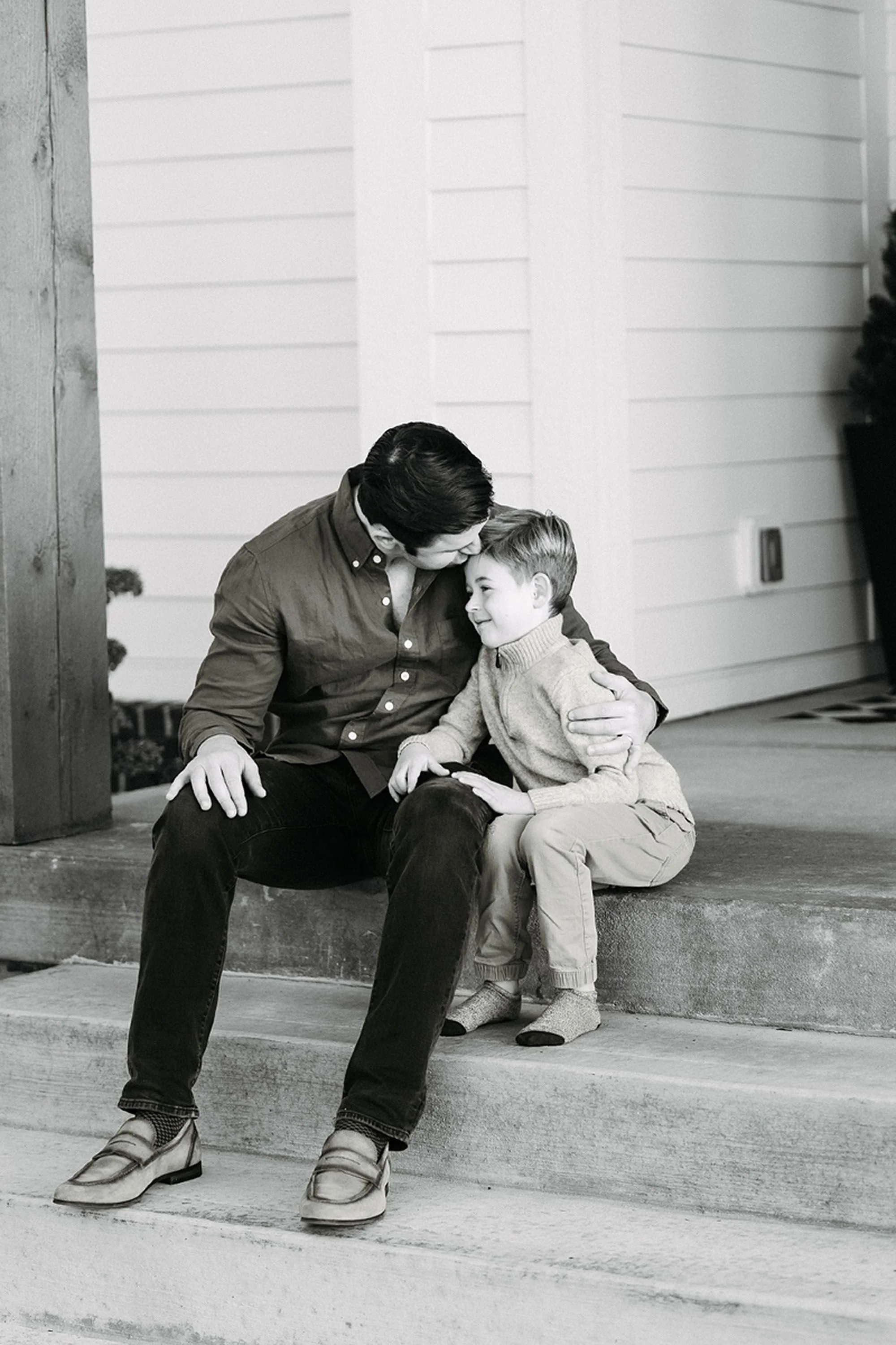 A father gently embracing his son on the front steps, capturing a tender father-son moment in a timeless black-and-white setting for at-home Christmas pictures in Chatham Hills.