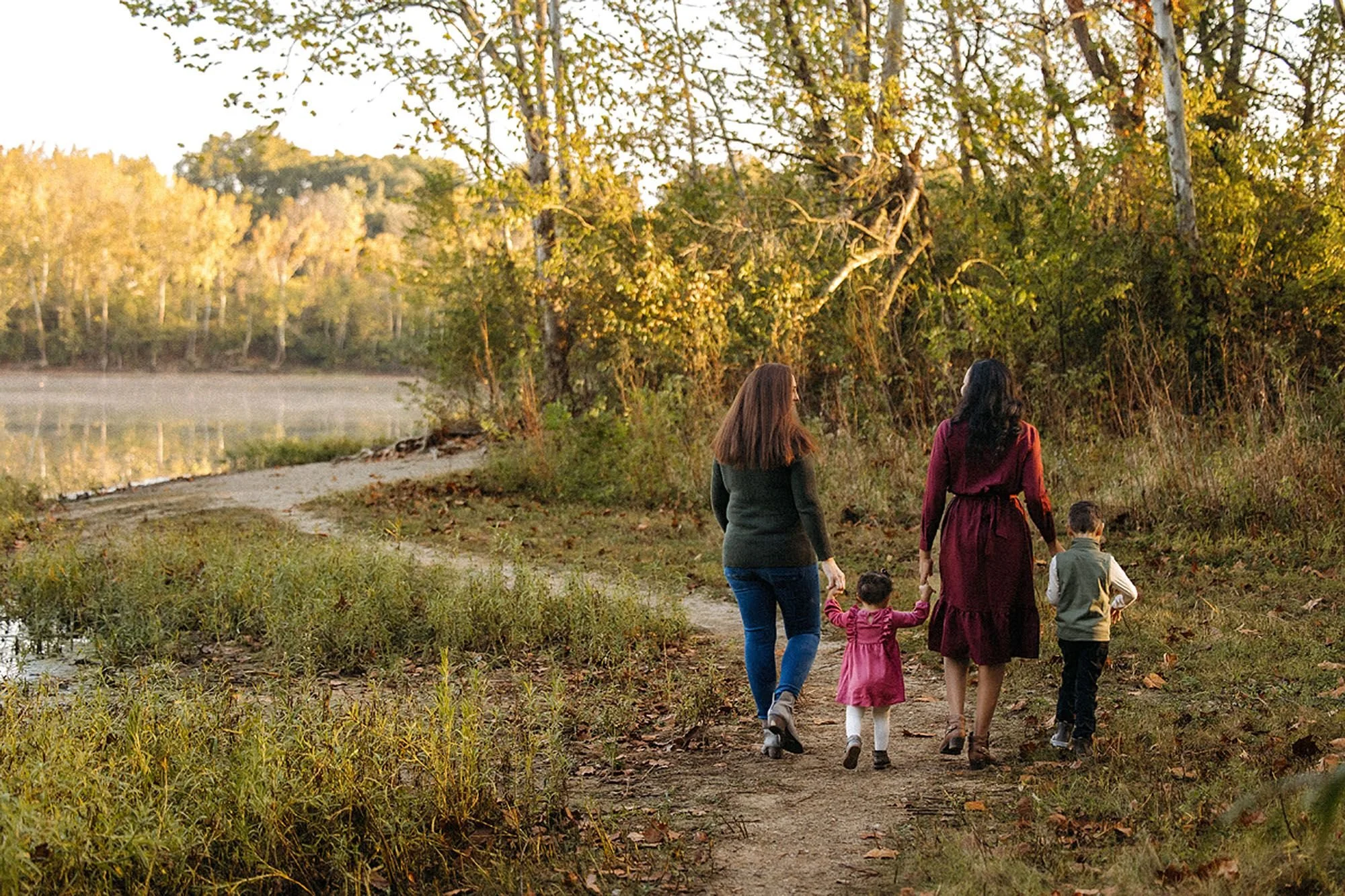 Family of four walking hand-in-hand along a wooded lakeside trail in autumn, seen from behind.